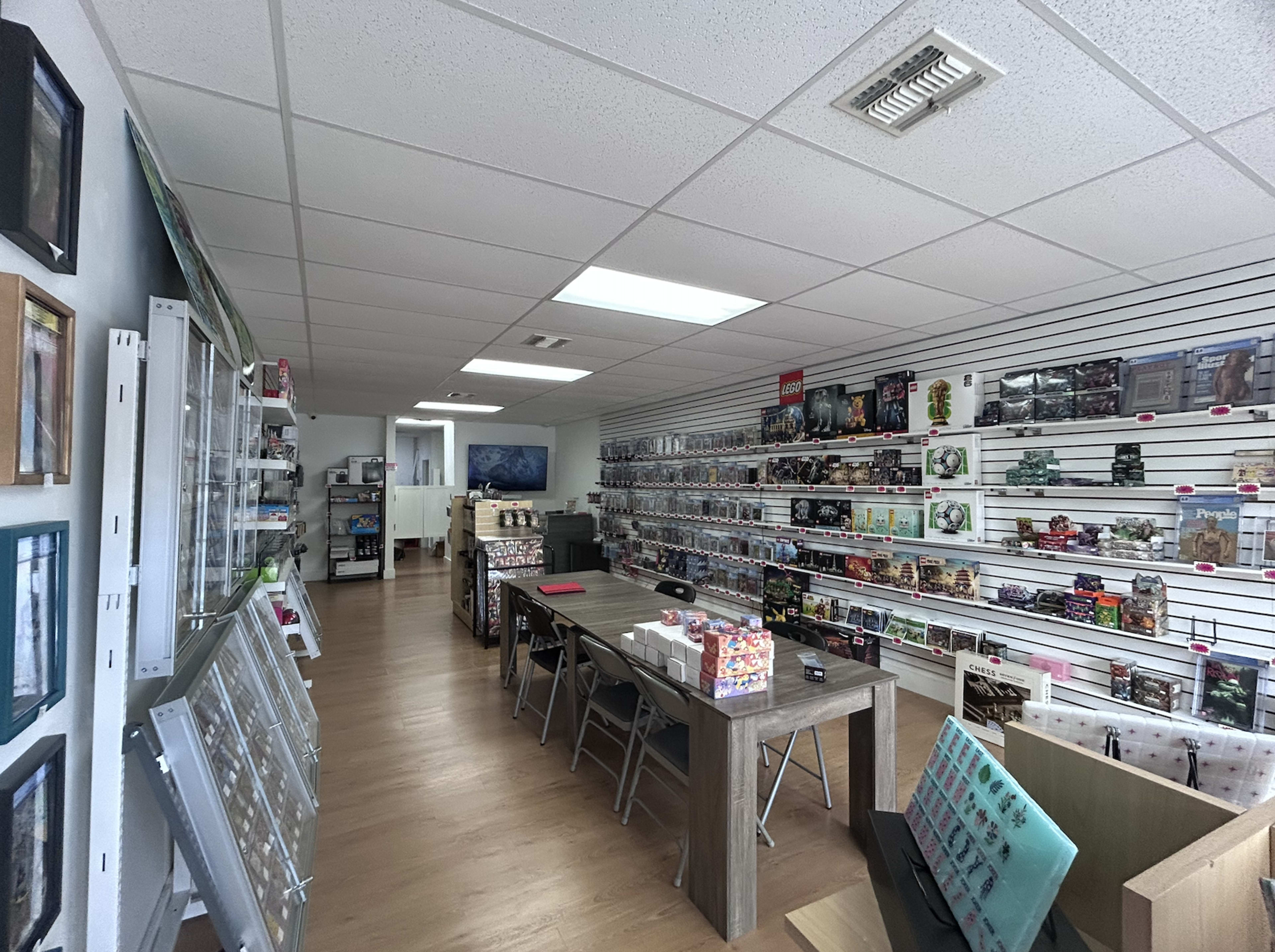 The image shows the interior of a retail store featuring neatly organized shelves displaying various products, with a central table and chairs.