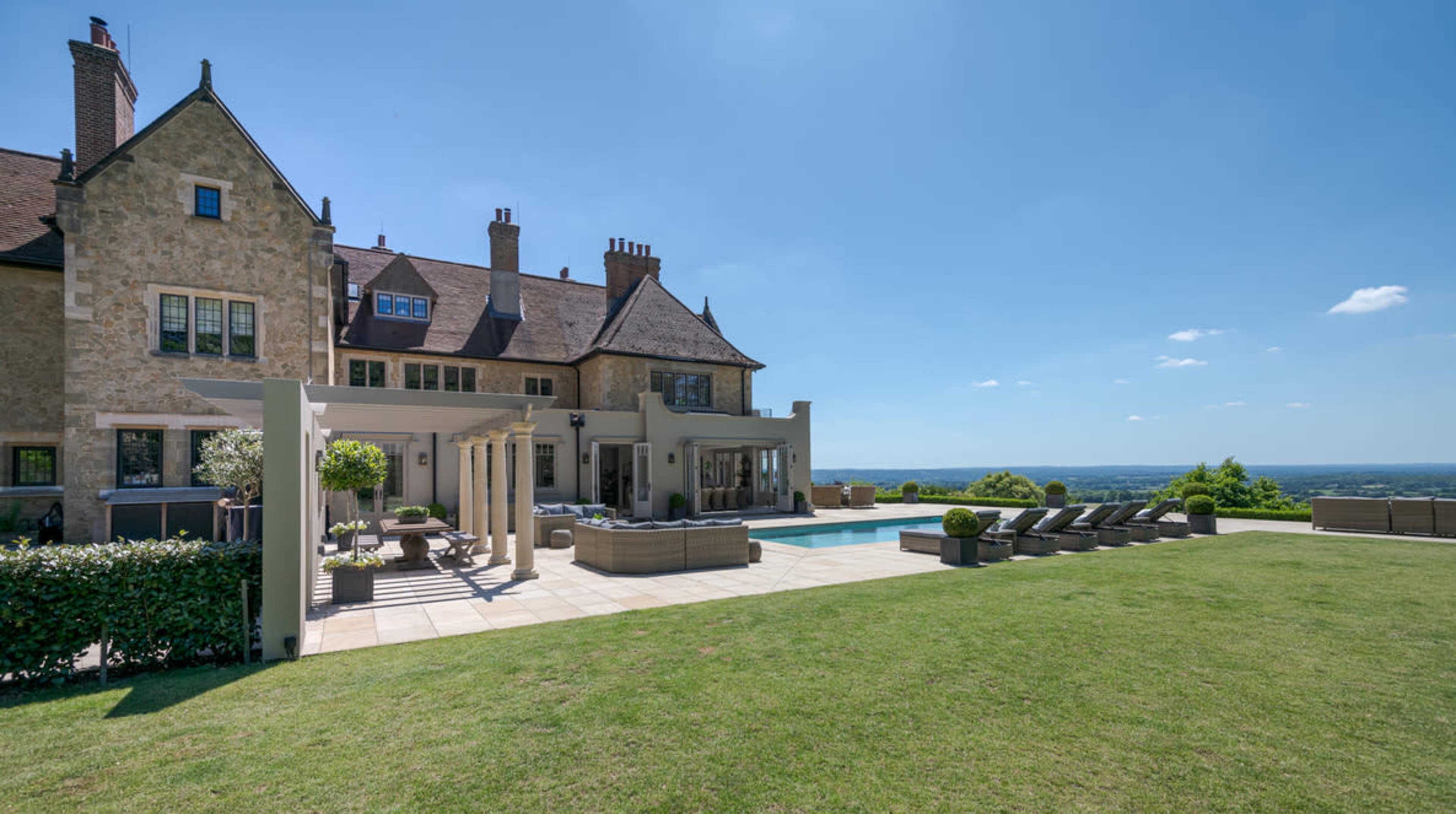 A large stone house with multiple chimneys overlooks a landscaped yard featuring a swimming pool and lounge chairs.