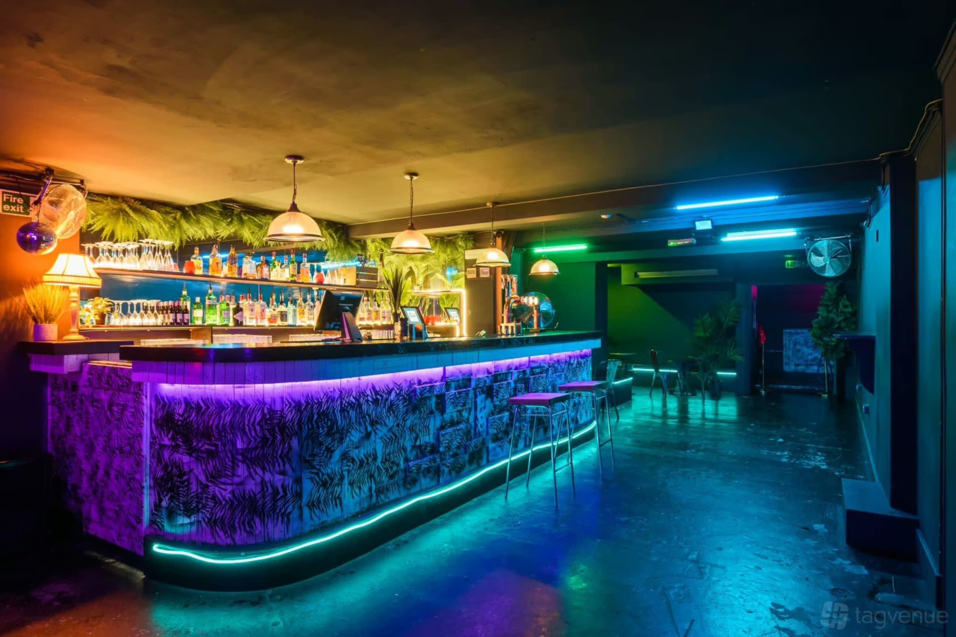A brightly lit bar featuring colorful neon lights, shelves stocked with various bottles, and a tropical-themed decor.