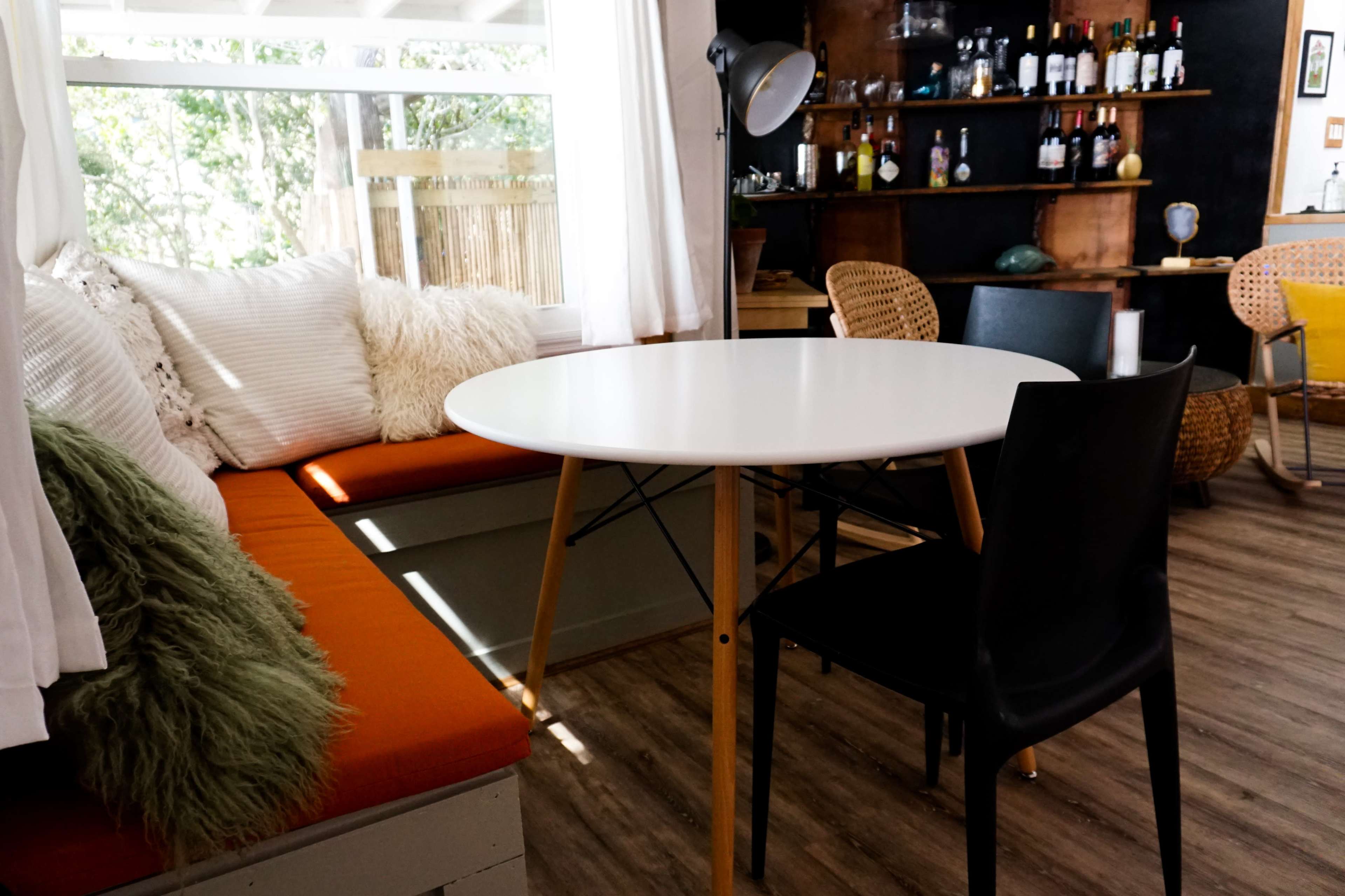 A round white table with wooden legs is positioned in a cozy seating area featuring a bench with orange cushions and a black chair.