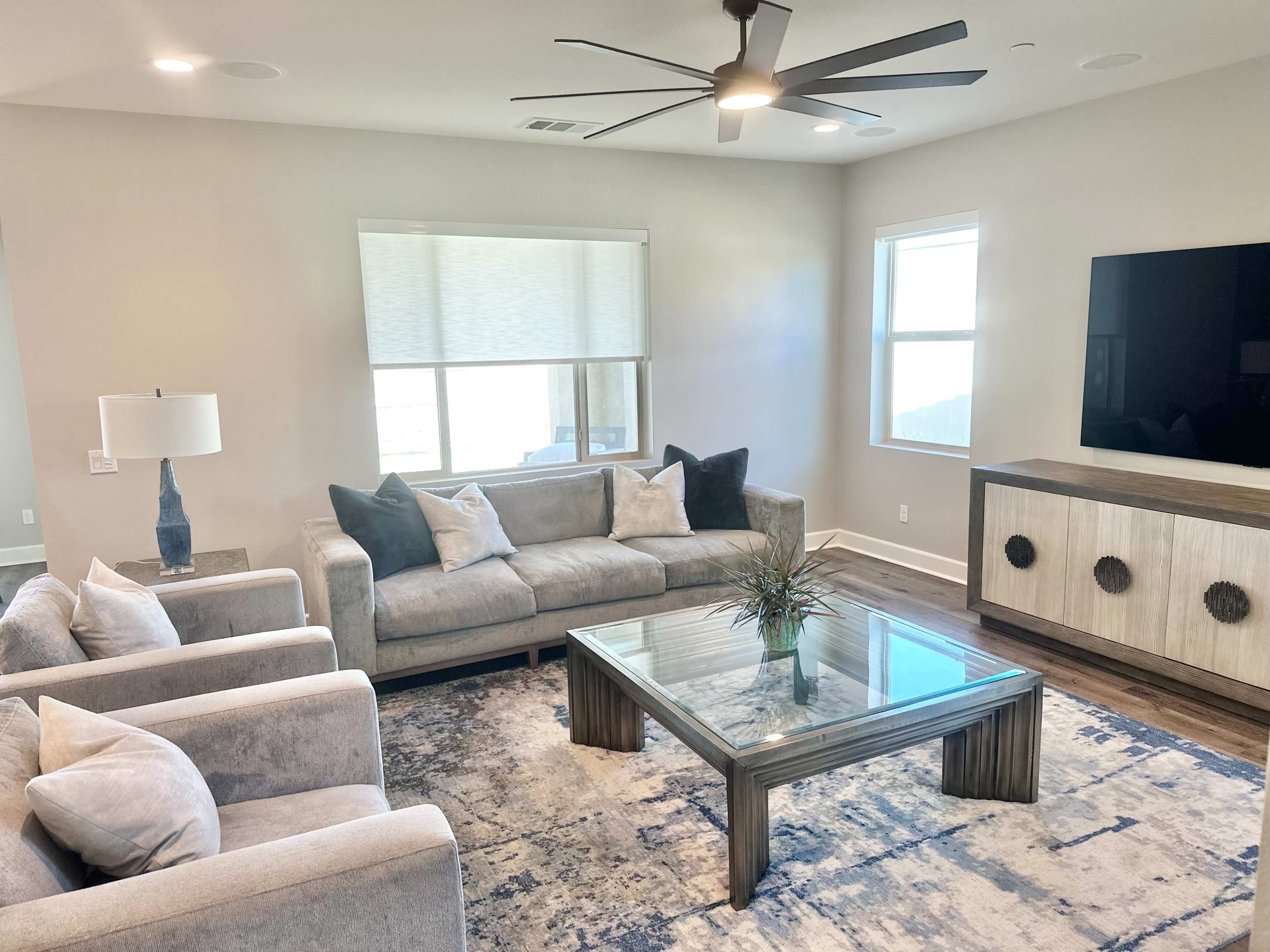 The image shows a modern living room with a gray sofa, two matching armchairs, a glass coffee table, and a television mounted on the wall.