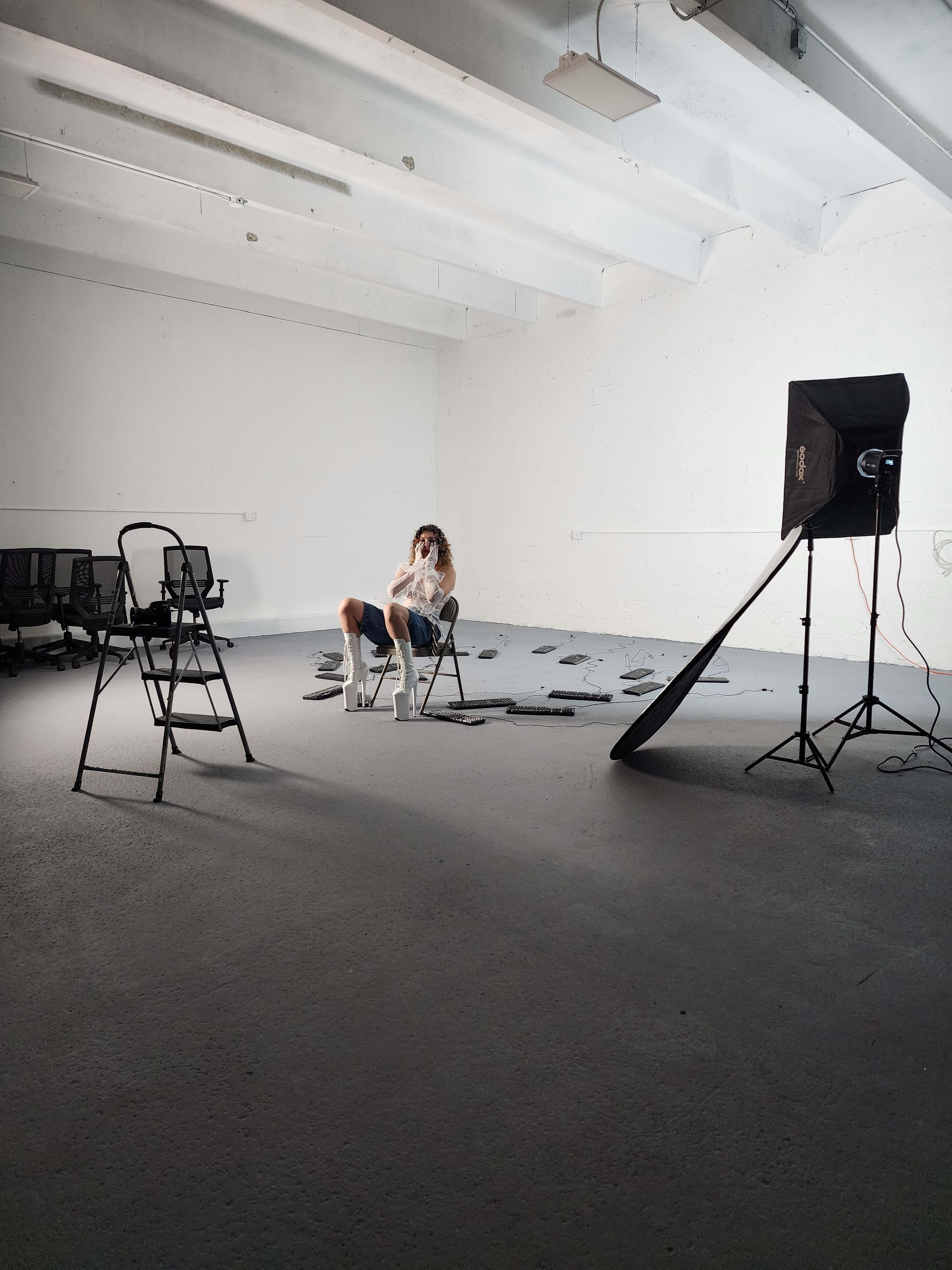 A person sits in a chair in an empty room with scattered objects on the floor and photography equipment set up nearby.