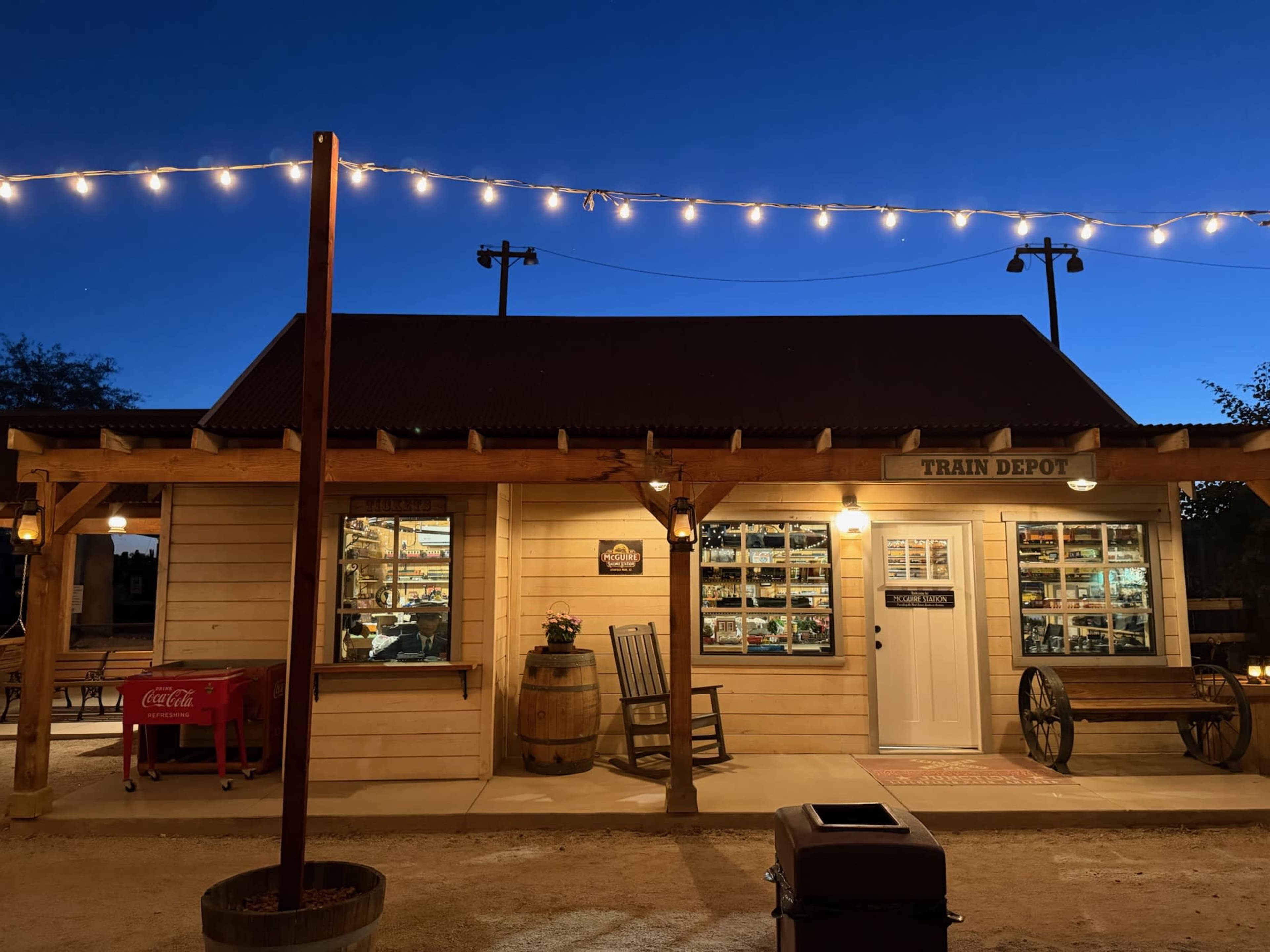 A wooden train depot building is illuminated by string lights, featuring large windows and a porch with seating.