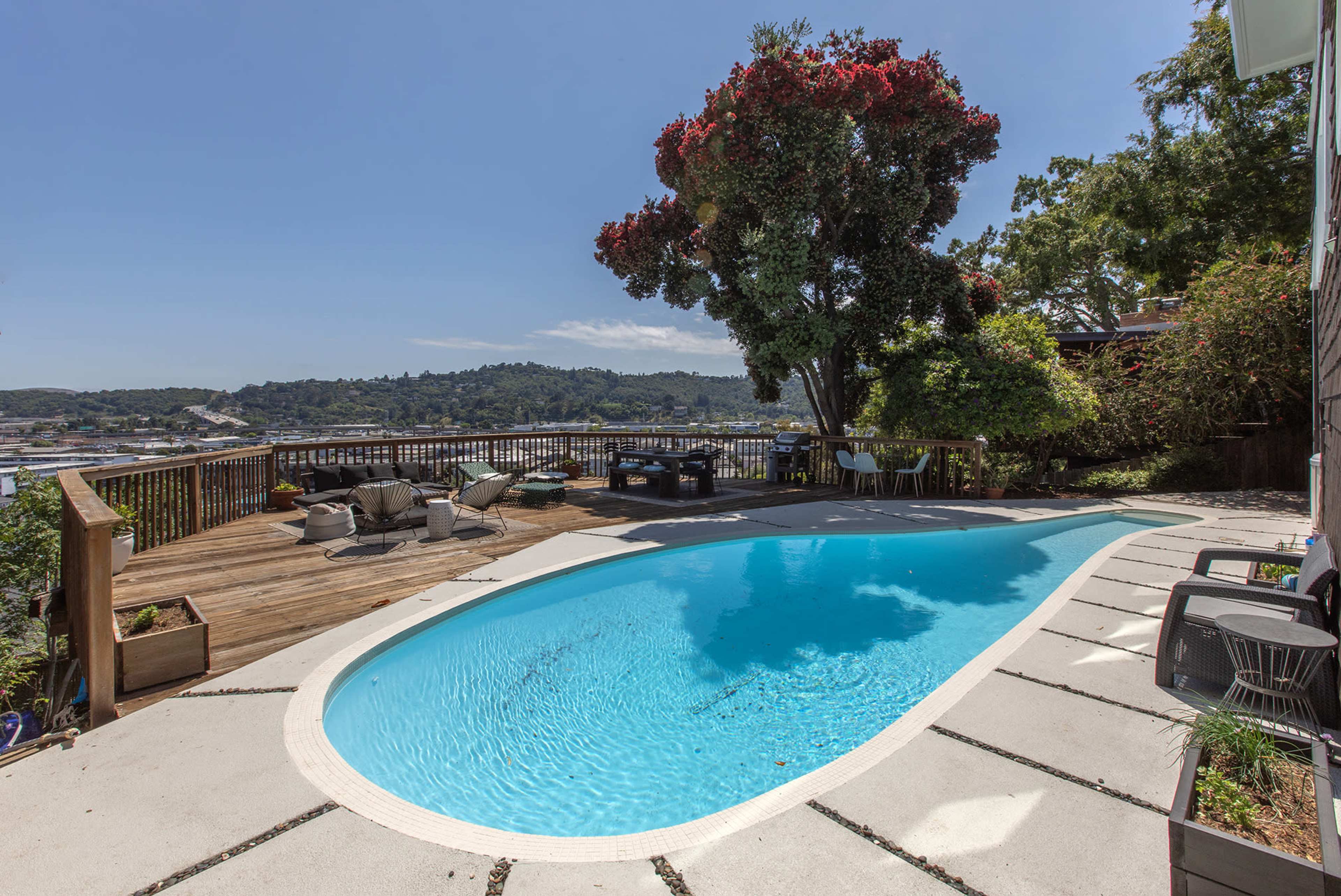 A rectangular swimming pool is set against a backdrop of trees and a scenic view, with a wooden deck and lounge chairs surrounding it.