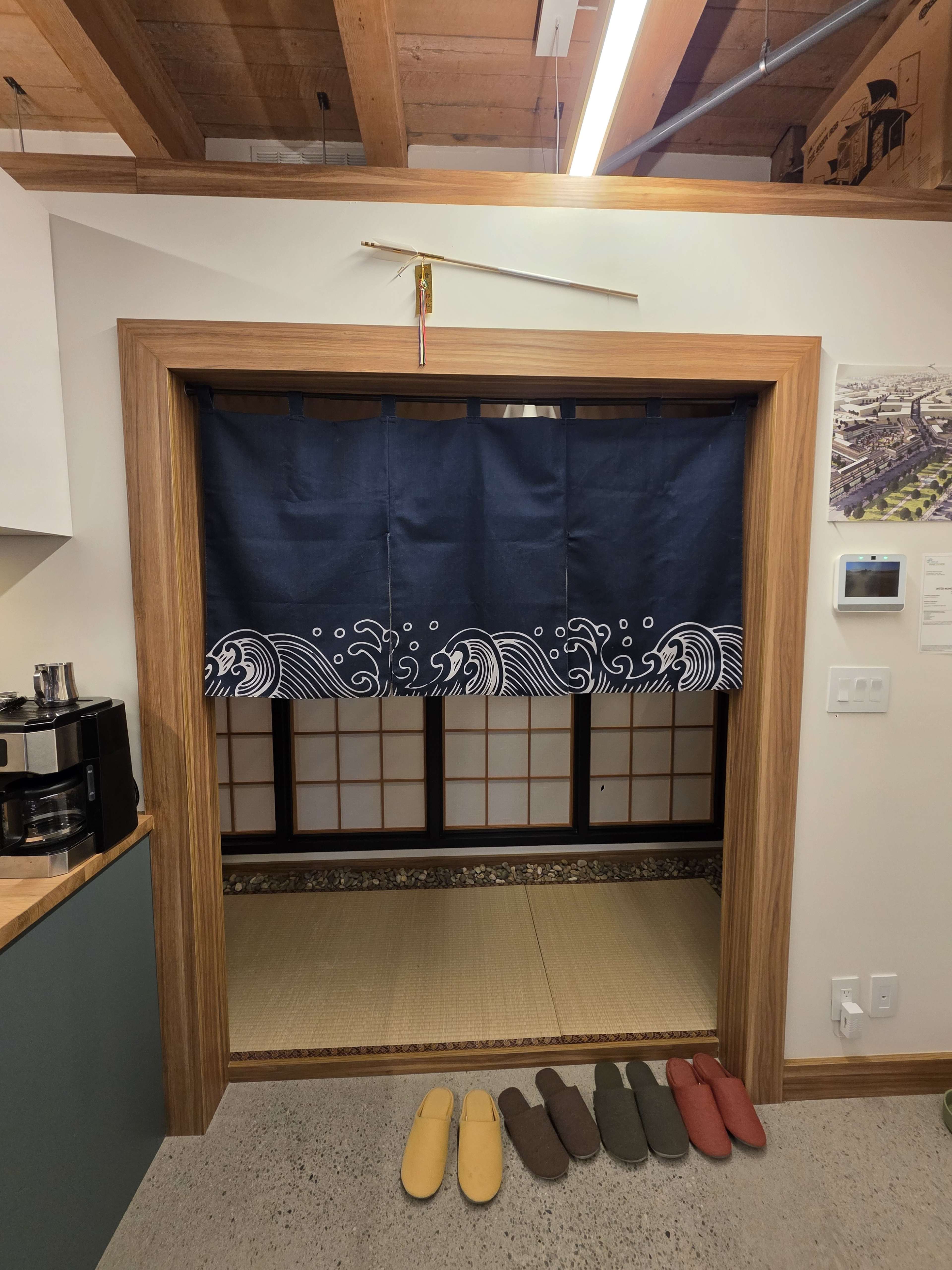 The image shows a wooden entrance framed by a blue fabric curtain featuring wave designs, with a tatami floor visible behind it and several pairs of slippers placed in front.