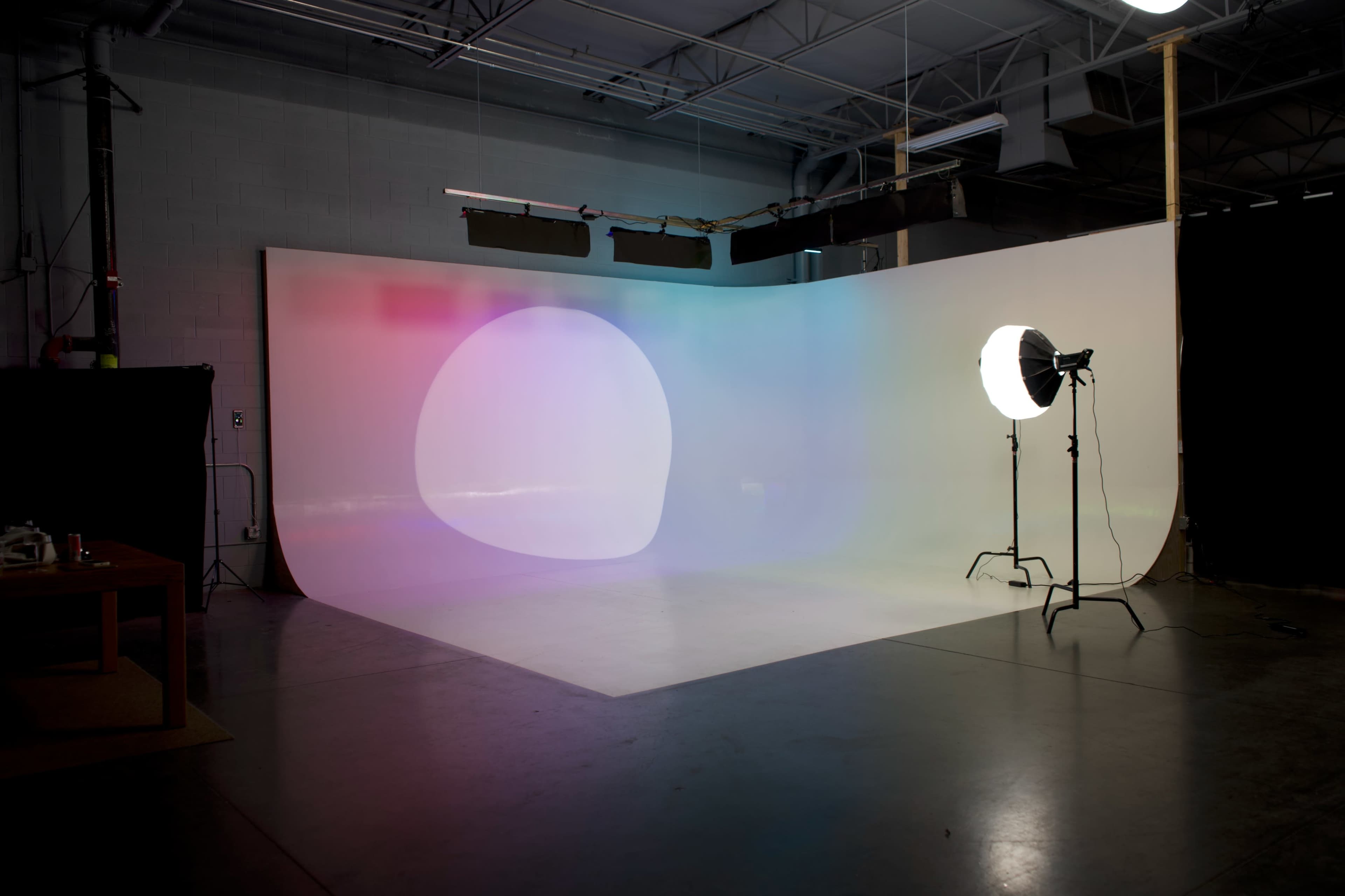 The image shows a sparse photo studio with a gradient backdrop and a large light source positioned on a stand.