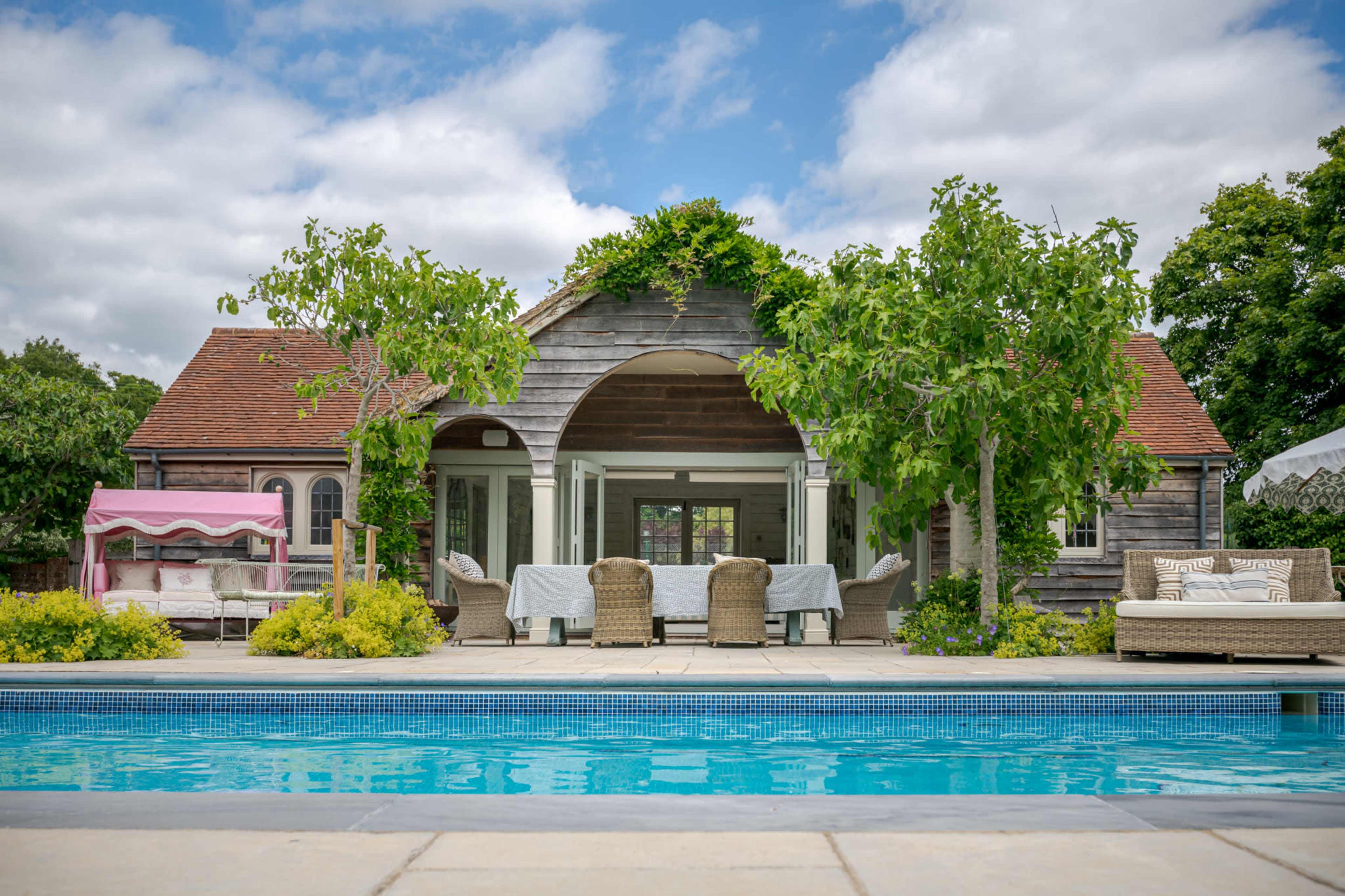 A modern pool area features a patio with wooden furniture and greenery, adjacent to a house with a wooden facade and large windows.