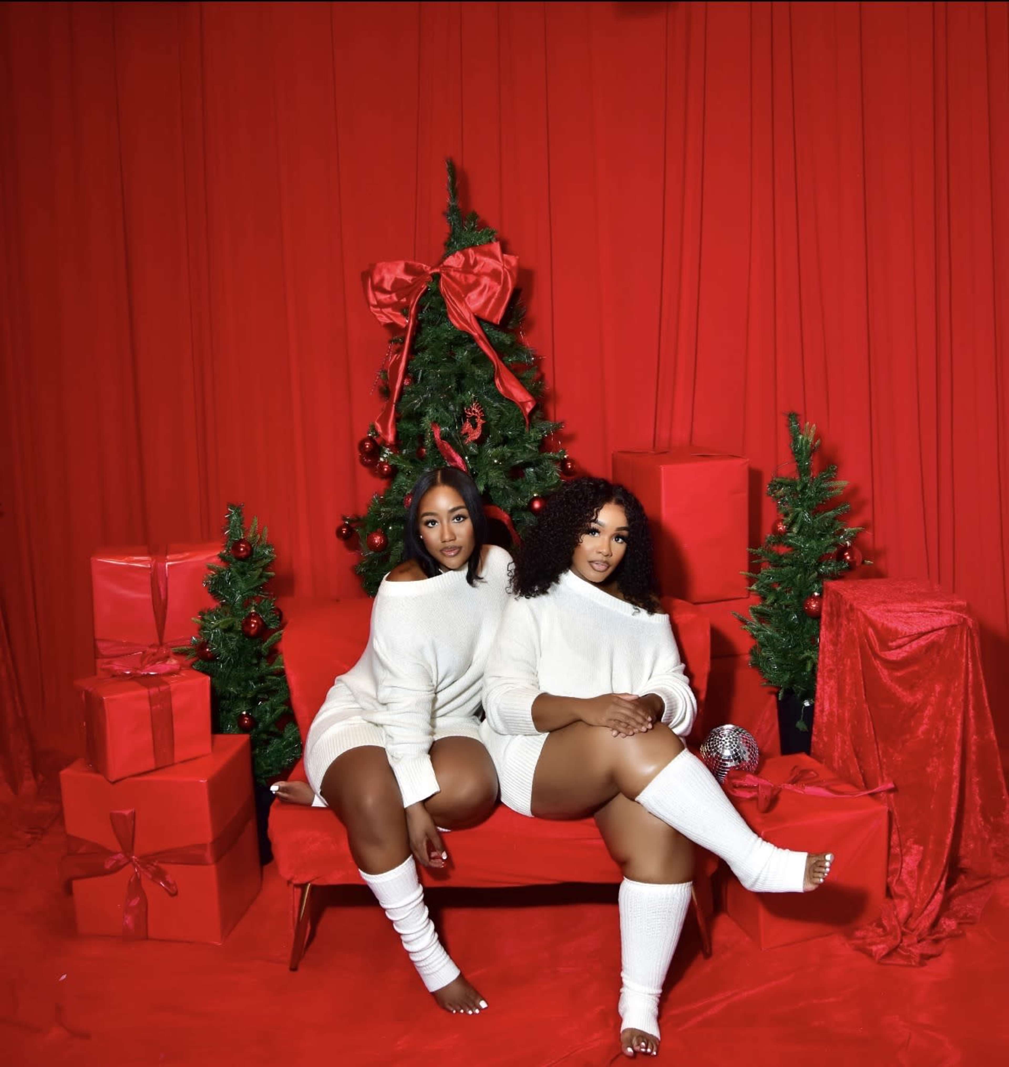 Two women in white outfits pose on a red couch surrounded by Christmas decorations, including a tree and wrapped gifts.
