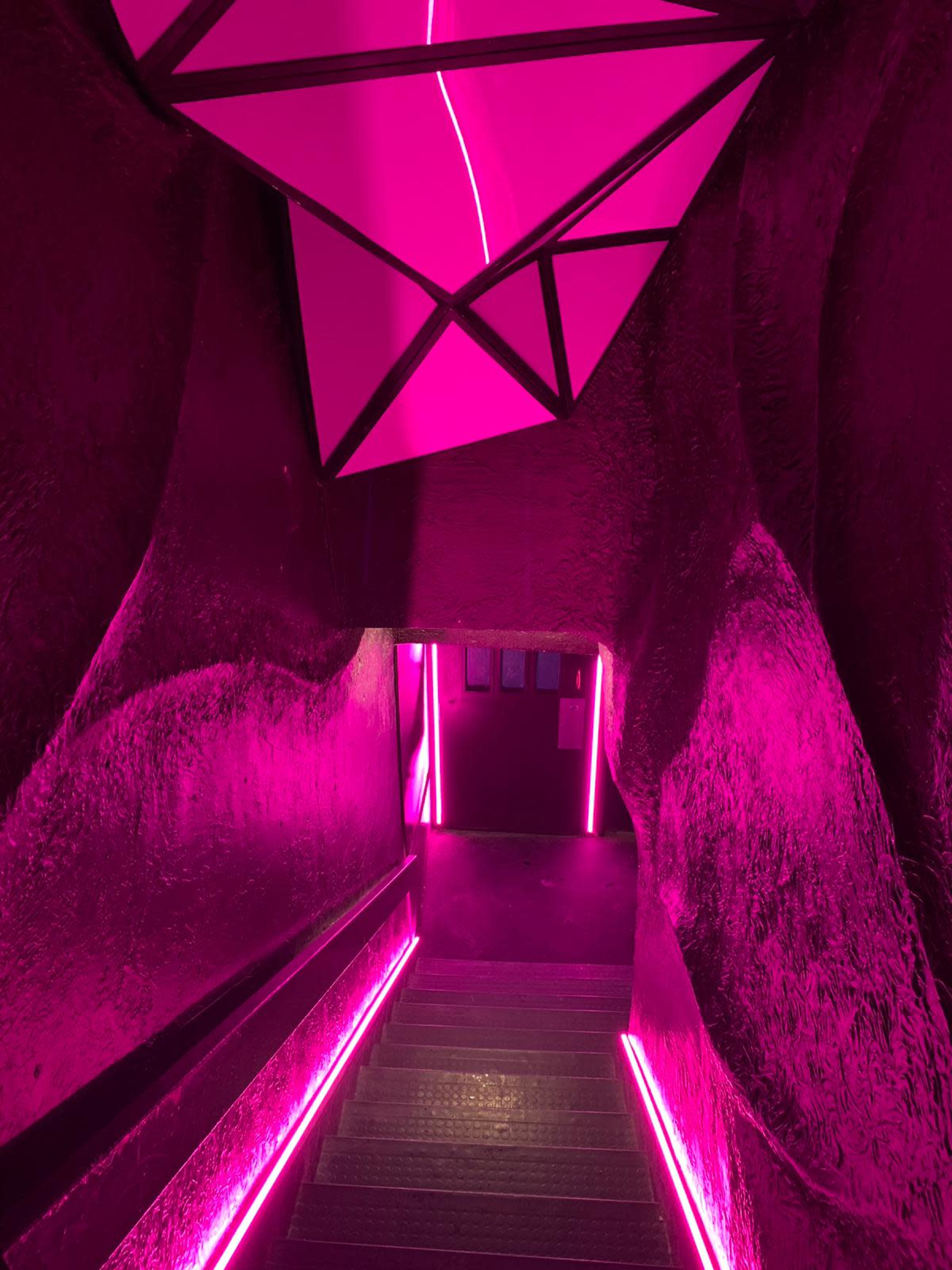 A staircase descends into a dimly lit space, illuminated by vibrant pink neon lights along the walls and ceiling.