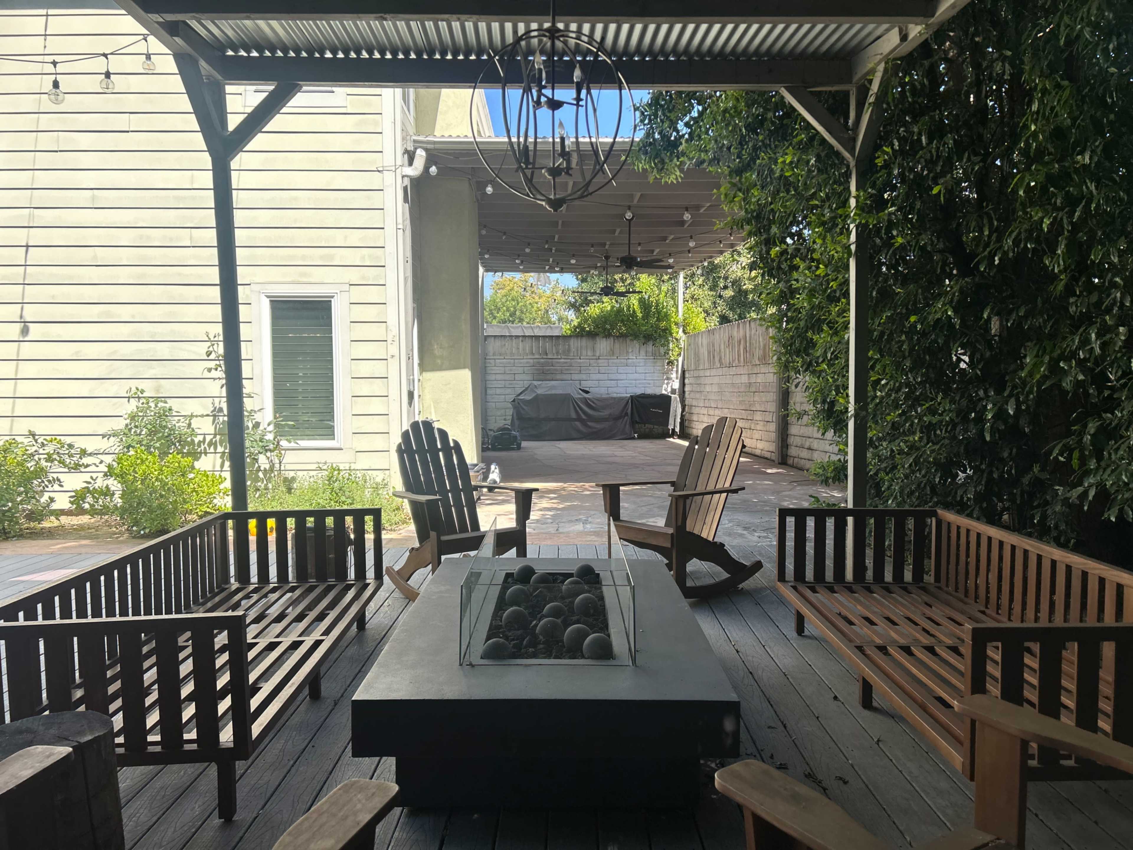 A patio area features a fire pit surrounded by wooden seating, with greenery and a covered space in the background.