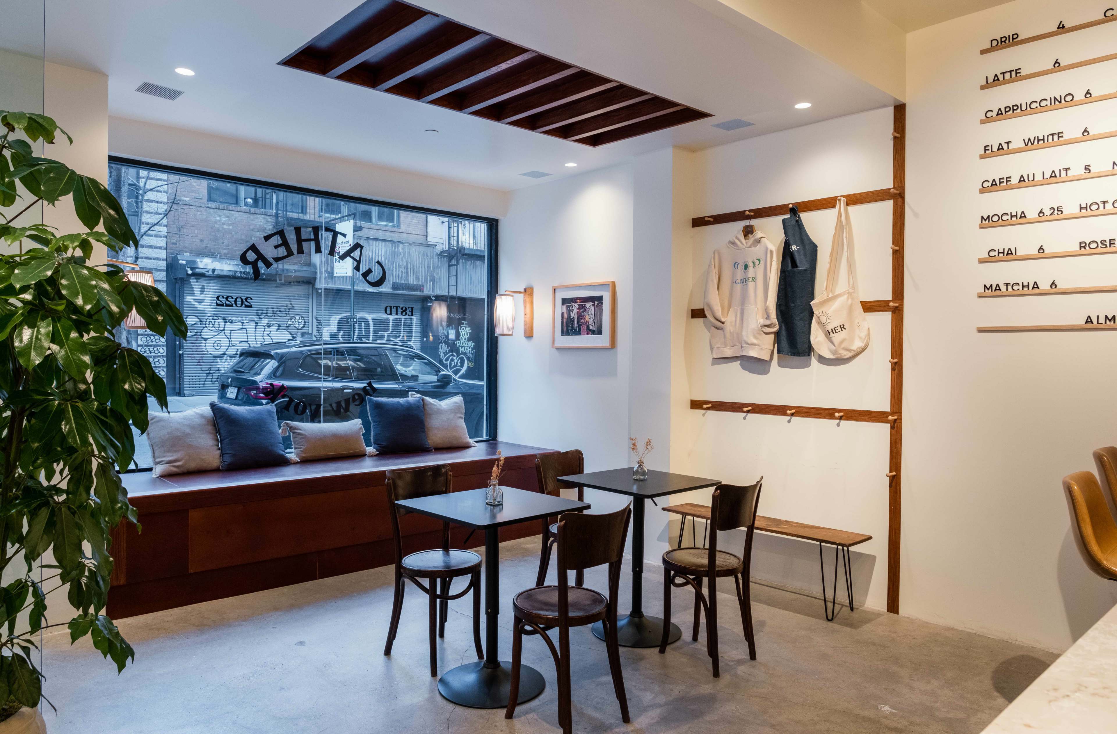 The image shows a minimalist café interior with a window view, a wooden bench with cushions, two small tables, and a display wall featuring menus and aprons.