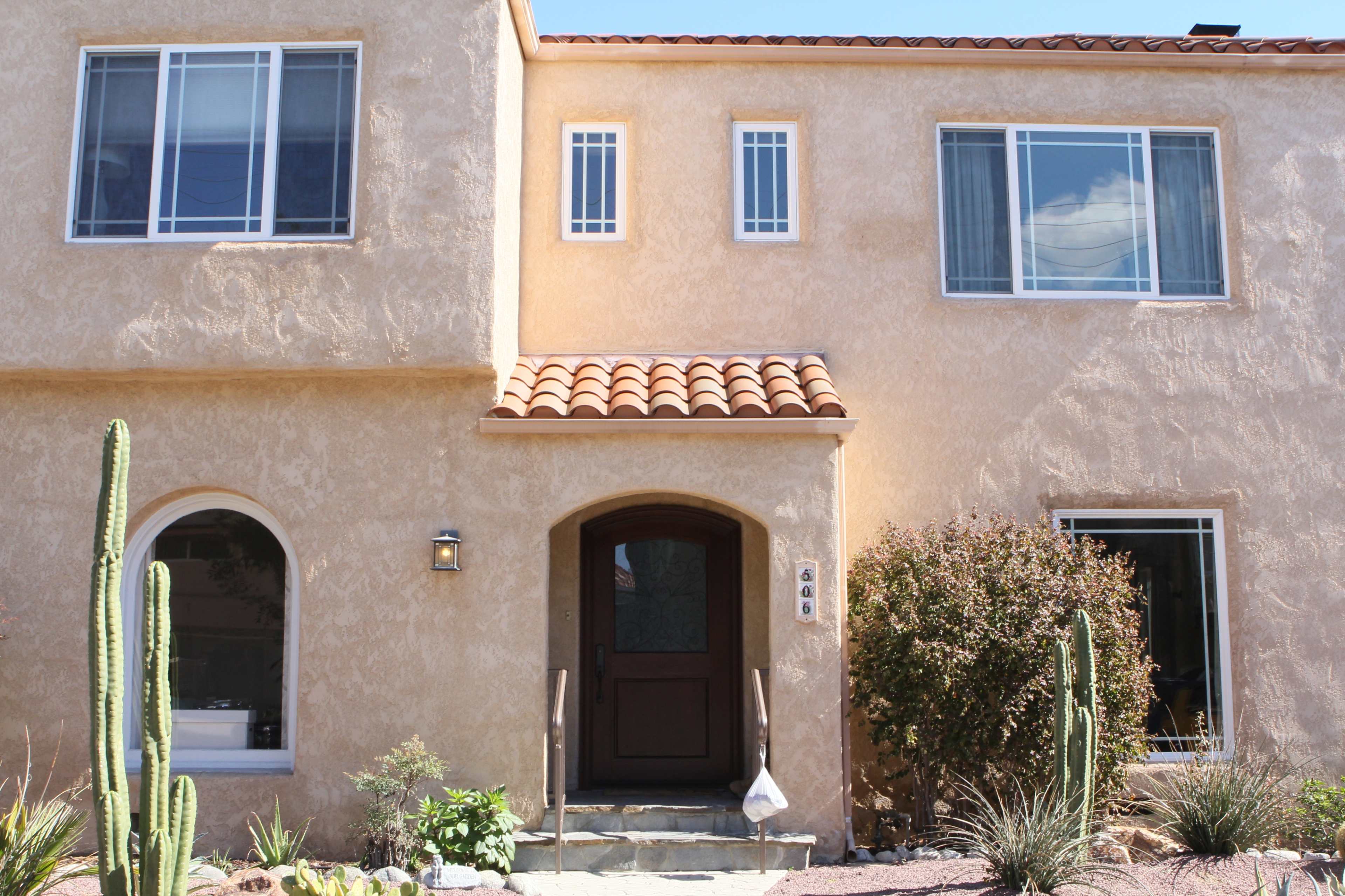 A two-story stucco house with a tiled roof, featuring multiple windows, a wooden front door, and desert landscaping with cacti.