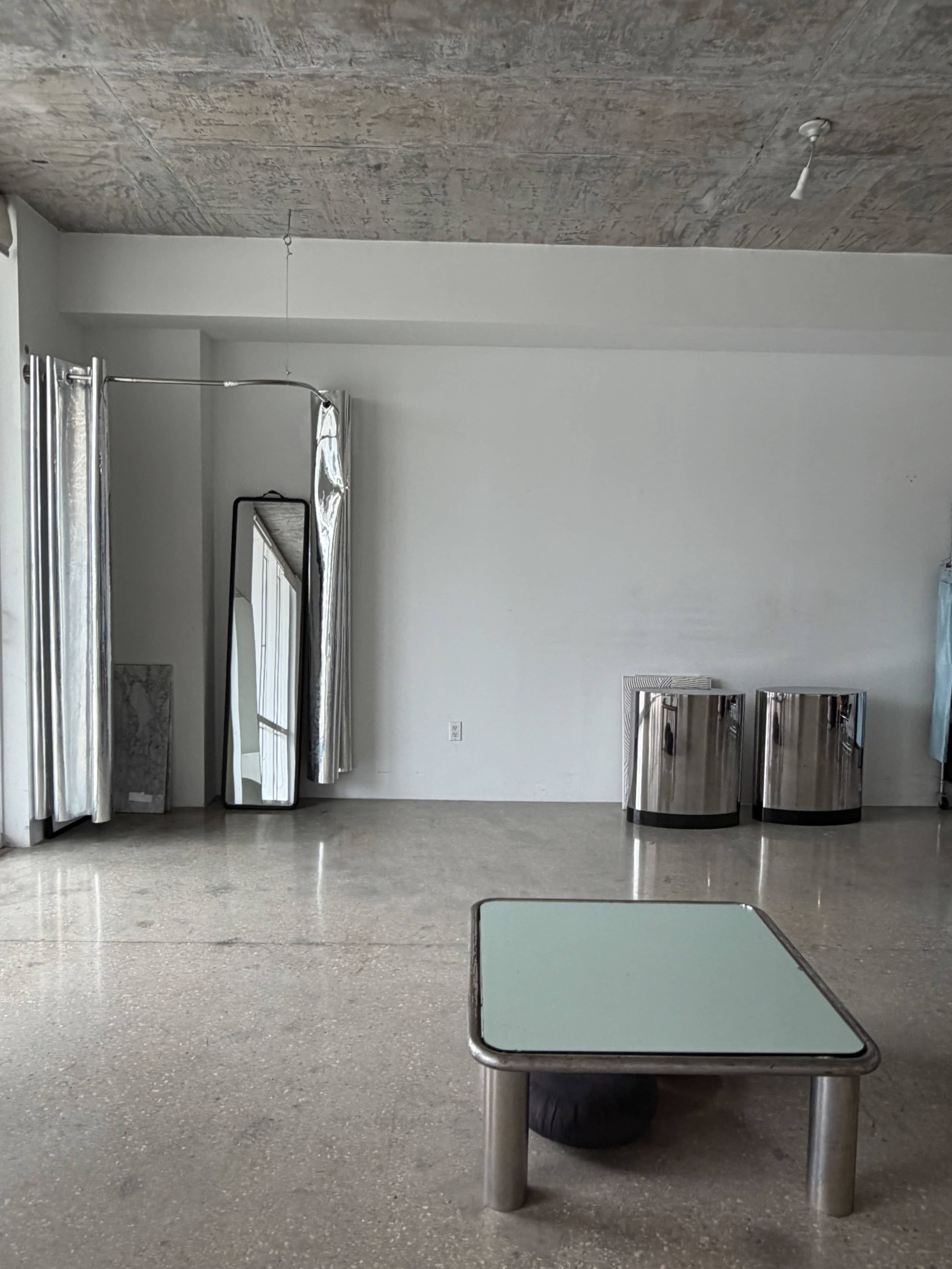 The image shows a minimalist interior with polished concrete flooring, a glass-top table, two cylindrical metal trash bins, a full-length mirror, and gray curtains.