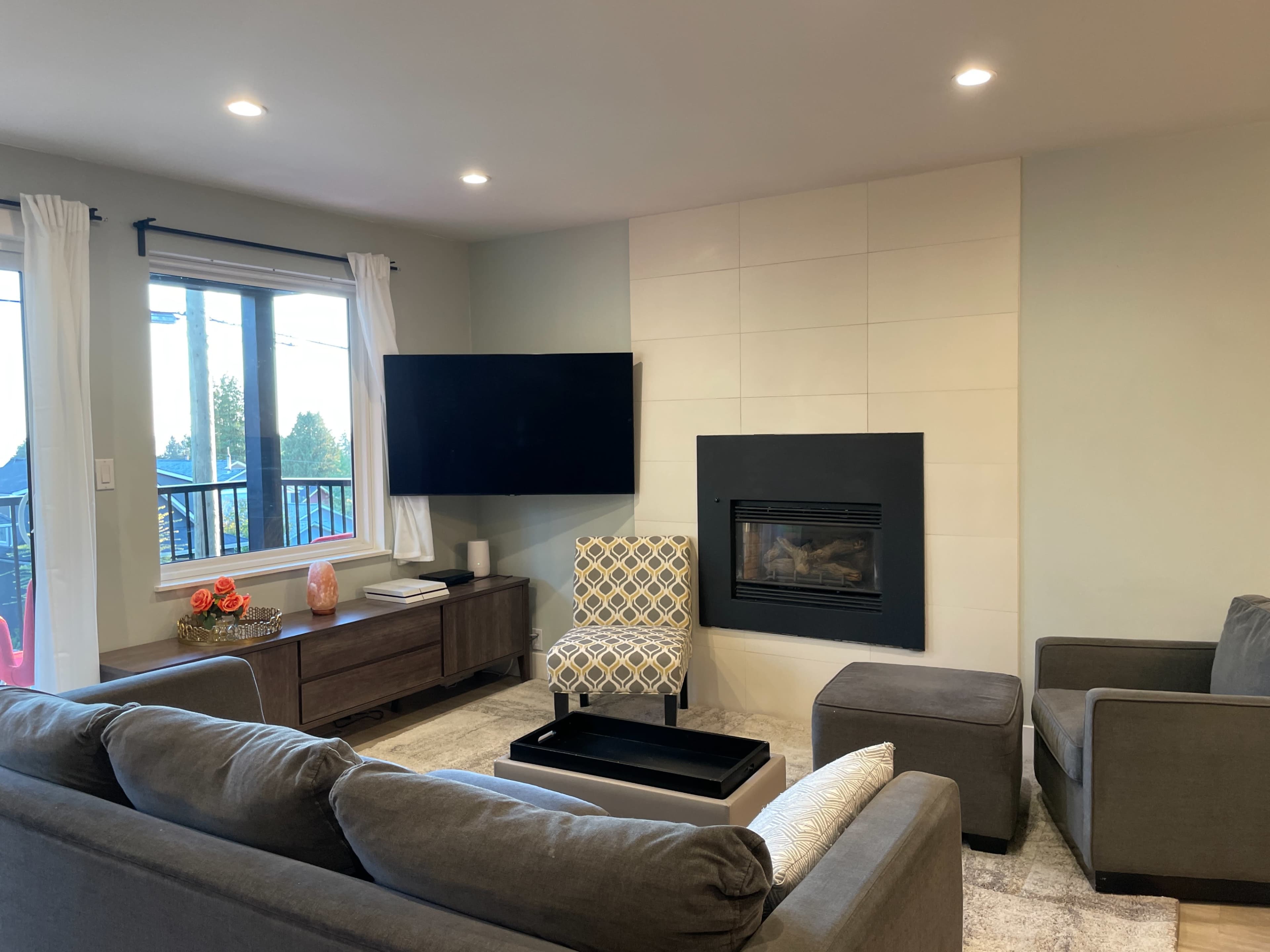 A living room with a television mounted on the wall, a fireplace, a gray sofa set, and large windows allowing natural light to enter.