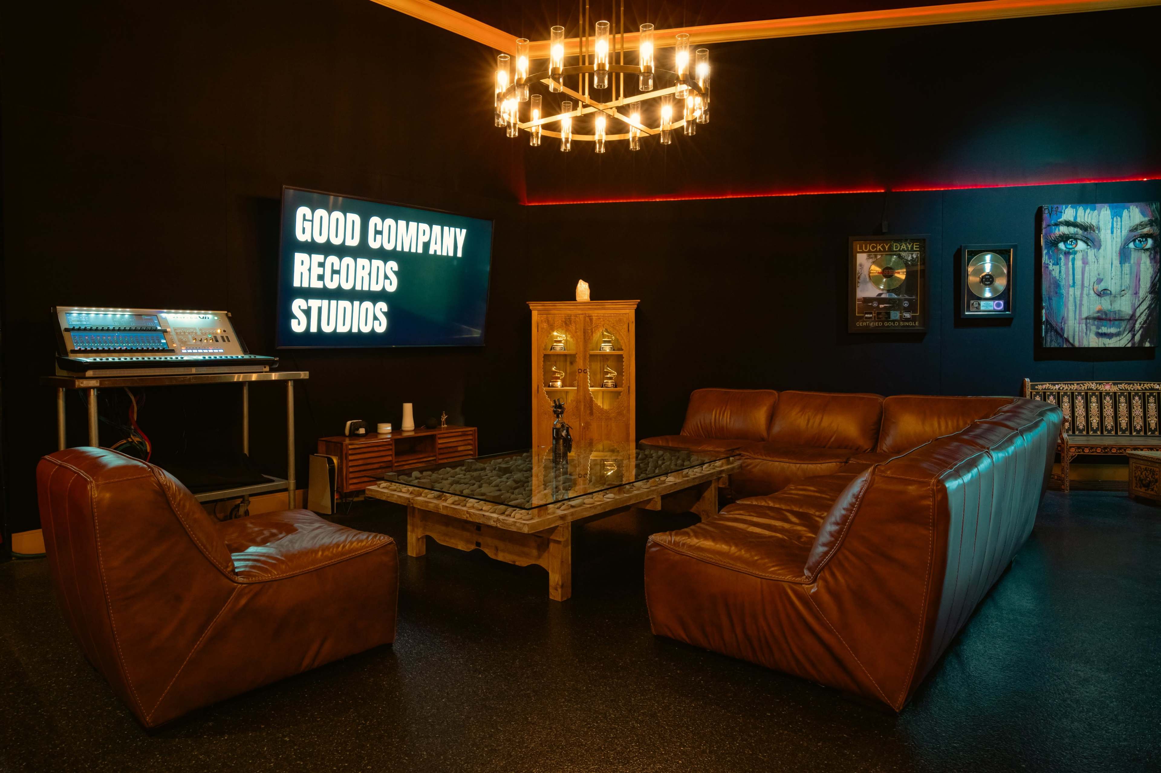 A recording studio lounge area featuring a leather sofa set, a glass-top coffee table, a television displaying "GOOD COMPANY RECORDS STUDIOS," and various decor elements including framed music plaques and a chandelier.