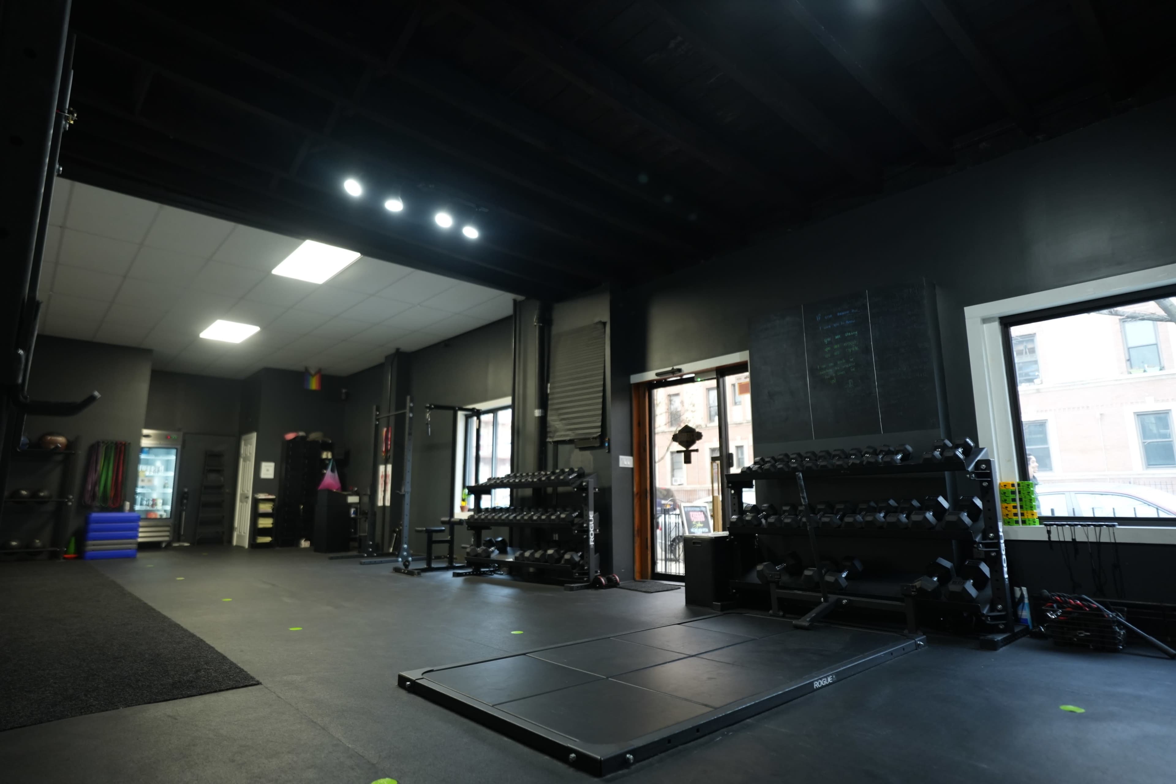 A gym interior featuring weightlifting equipment, a designated lifting platform, and large windows letting in natural light.