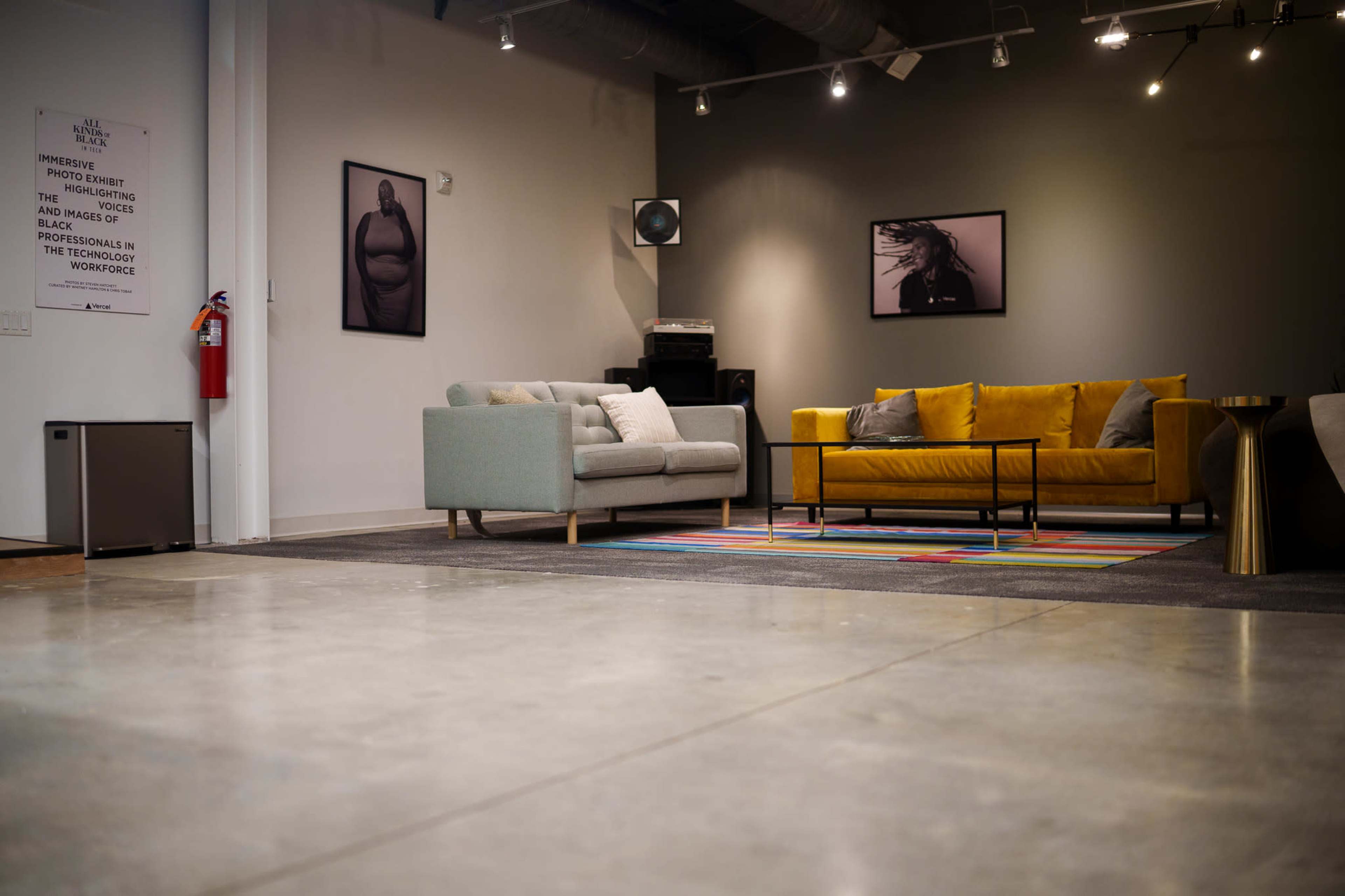 The image shows a modern interior space with a yellow couch, a light gray sofa, a colorful area rug, and two framed photographs on the walls.