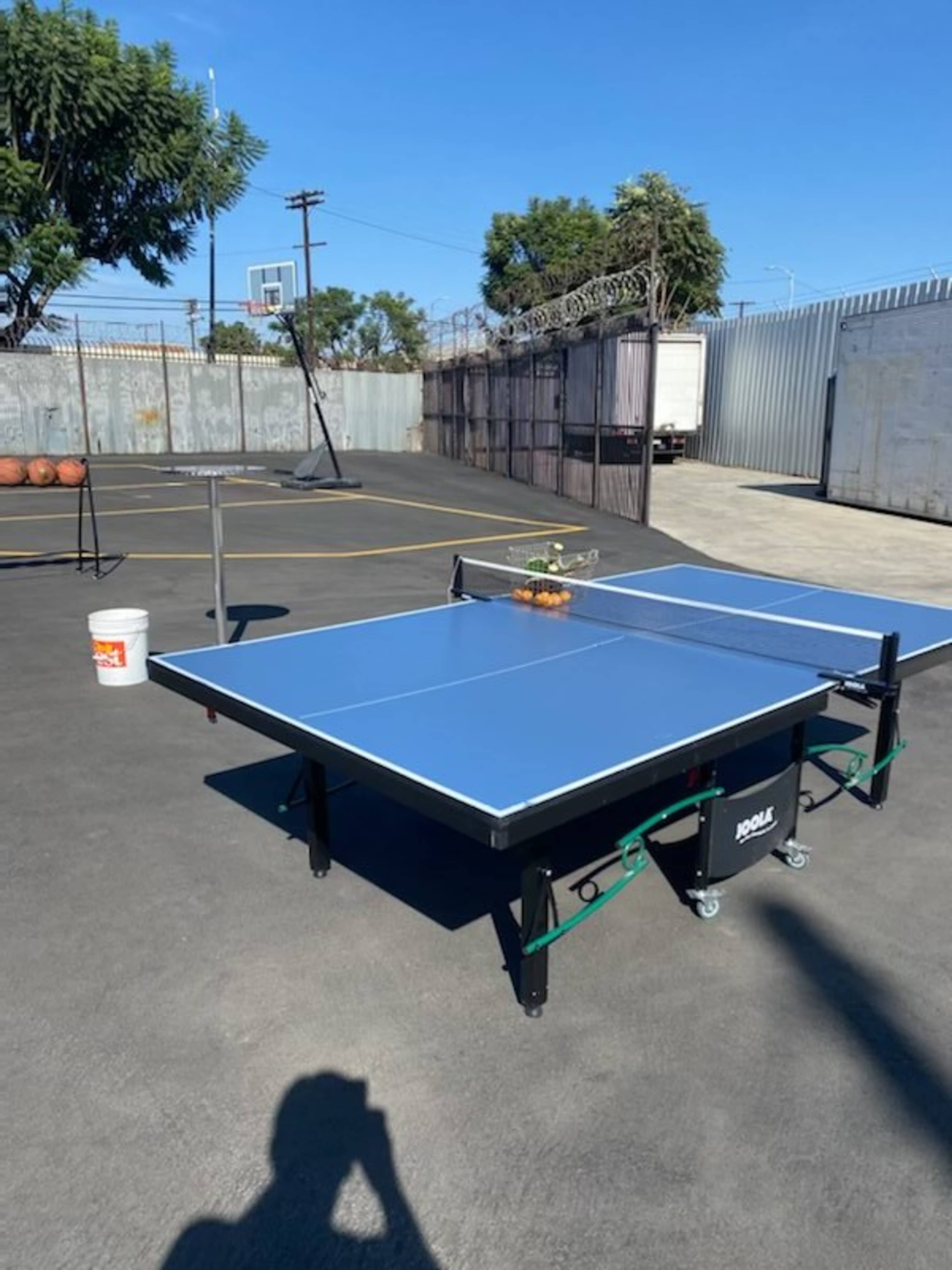 A ping pong table is set up outdoors on a paved surface next to a basketball court and a fenced area.