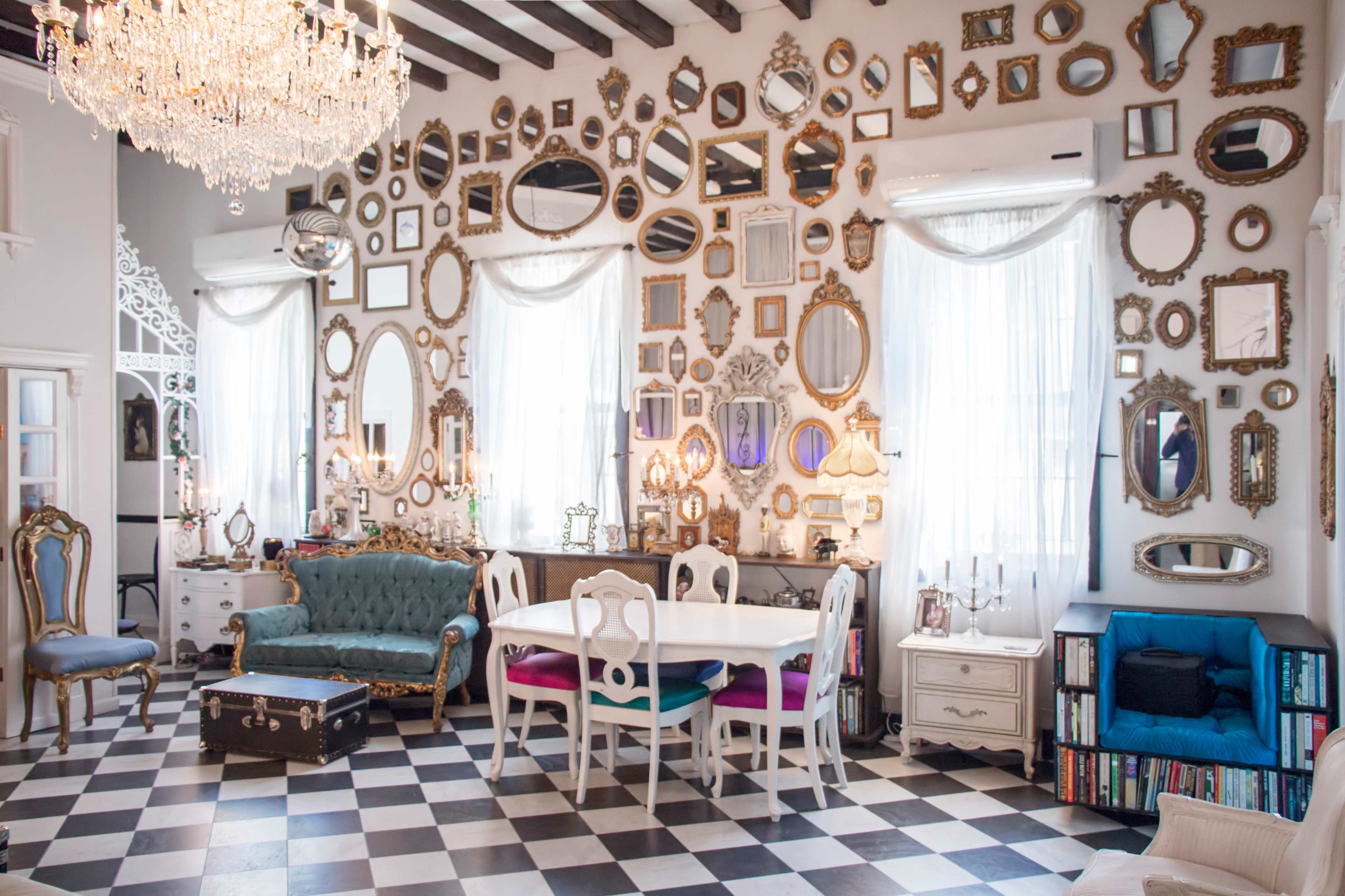 The room features a checkered floor with various mirrors hung on the walls, a chandelier above, and a mix of vintage furniture including a green couch and a white dining table.