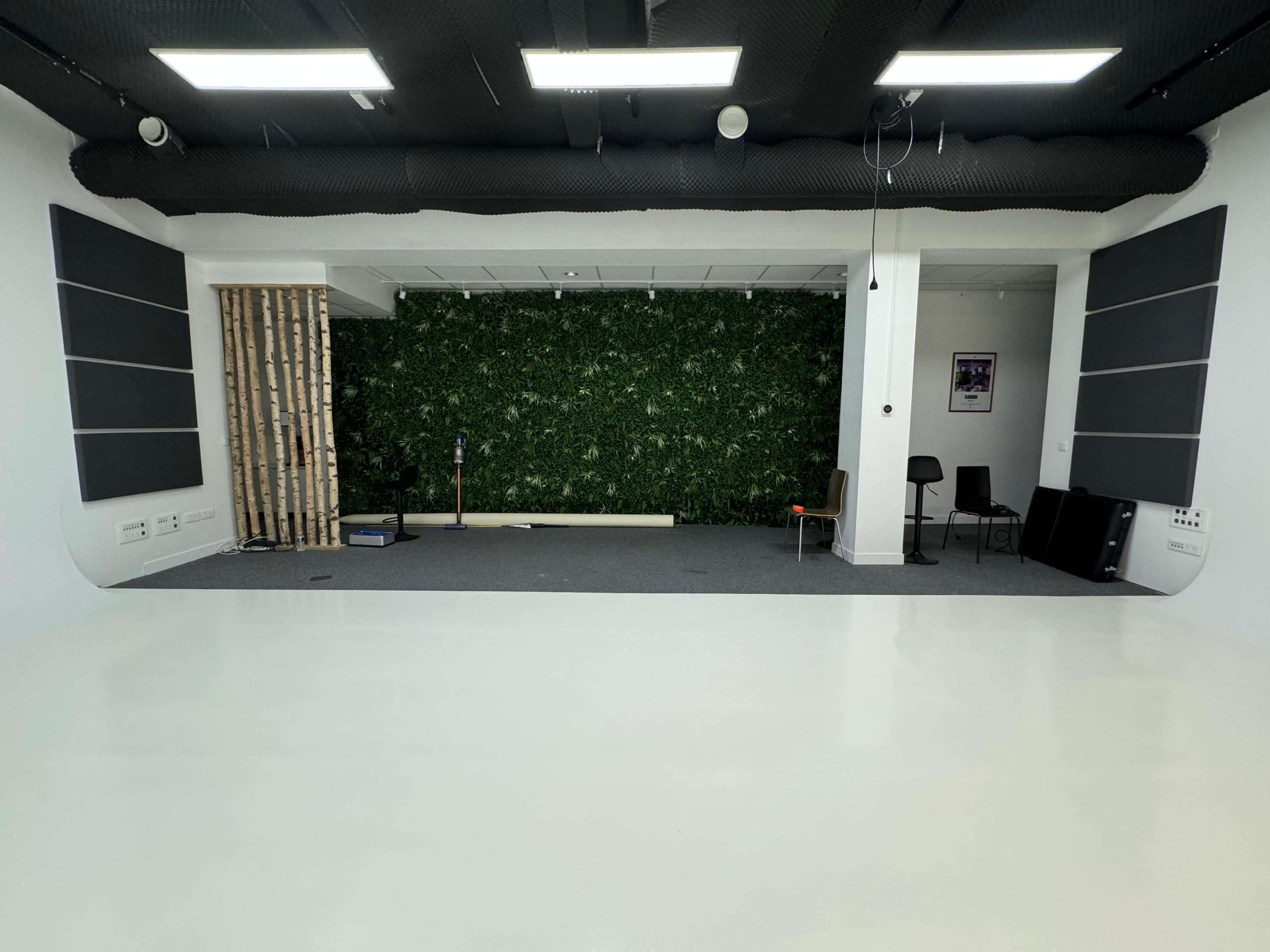 The image shows a spacious room with a white floor, acoustic panels on the walls, a green plant wall, and minimalist furniture arranged in a studio-like setting.