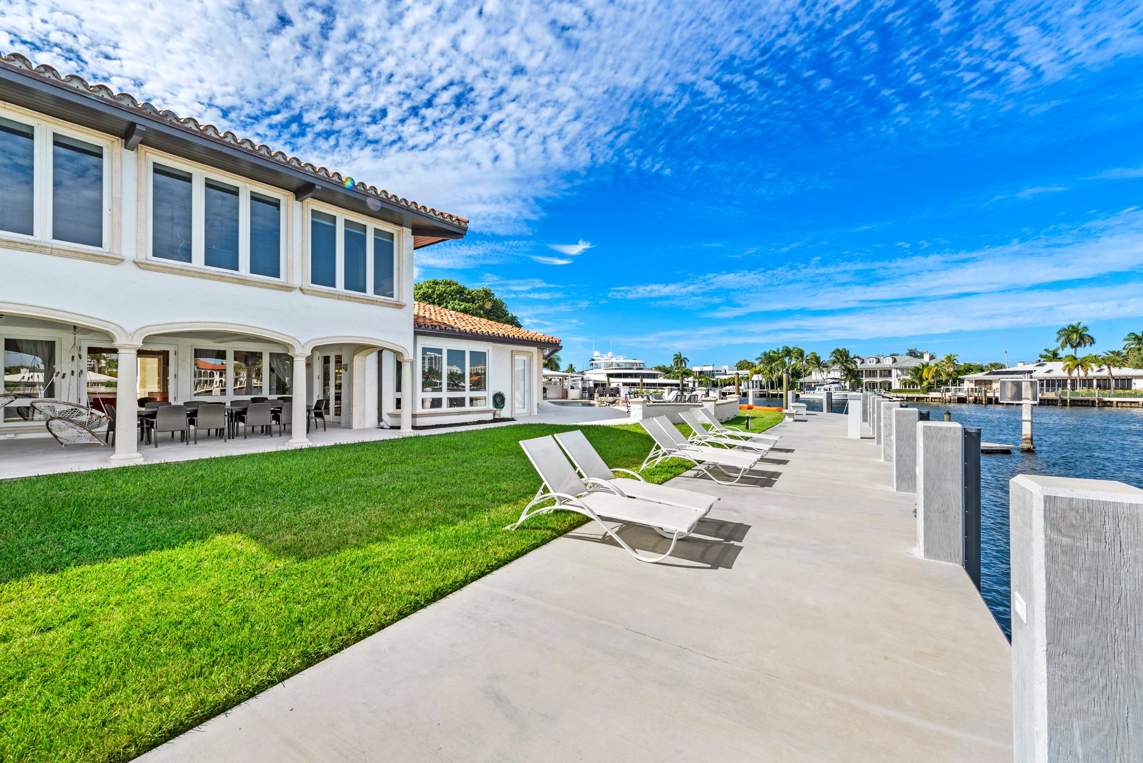 A waterfront property features a concrete path lined with lounge chairs next to a grassy area and a canal.