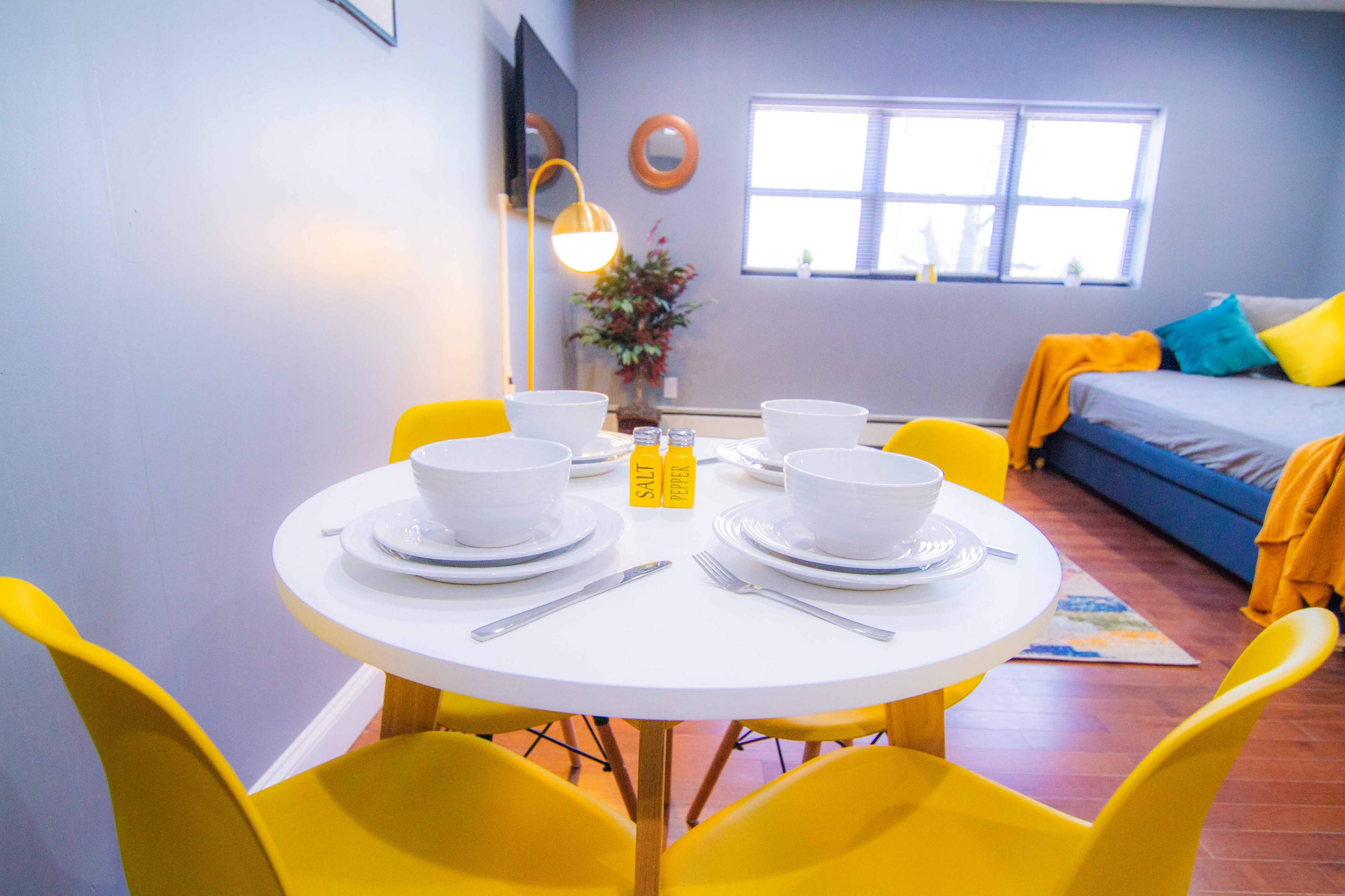 A round white table is set with bowls and utensils, surrounded by yellow chairs, in a room featuring a gray wall and a bed in the background.