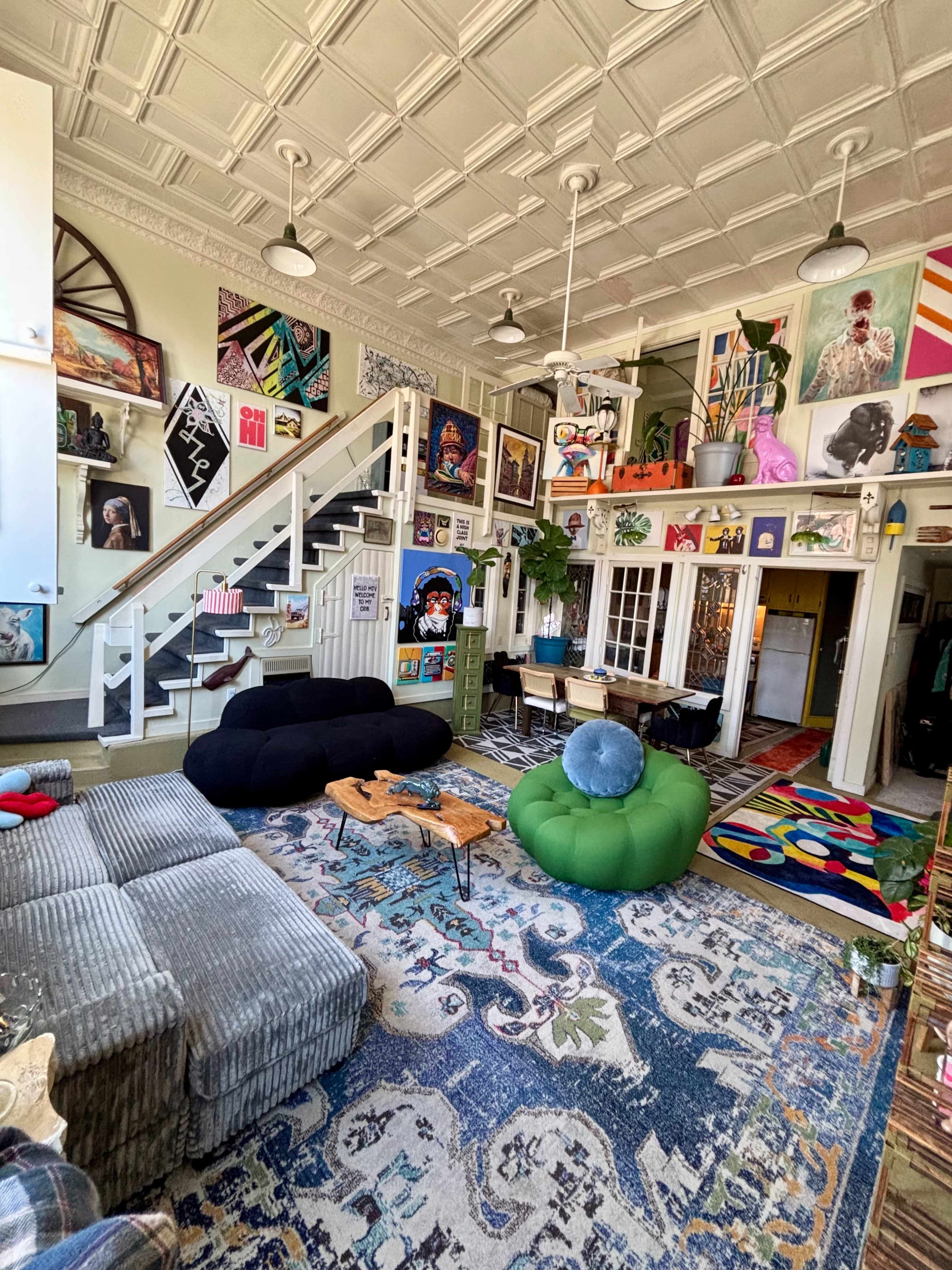 The image shows a spacious living room with a variety of art on the walls, a staircase leading to an upper level, and colorful furniture including bean bags and a patterned rug.