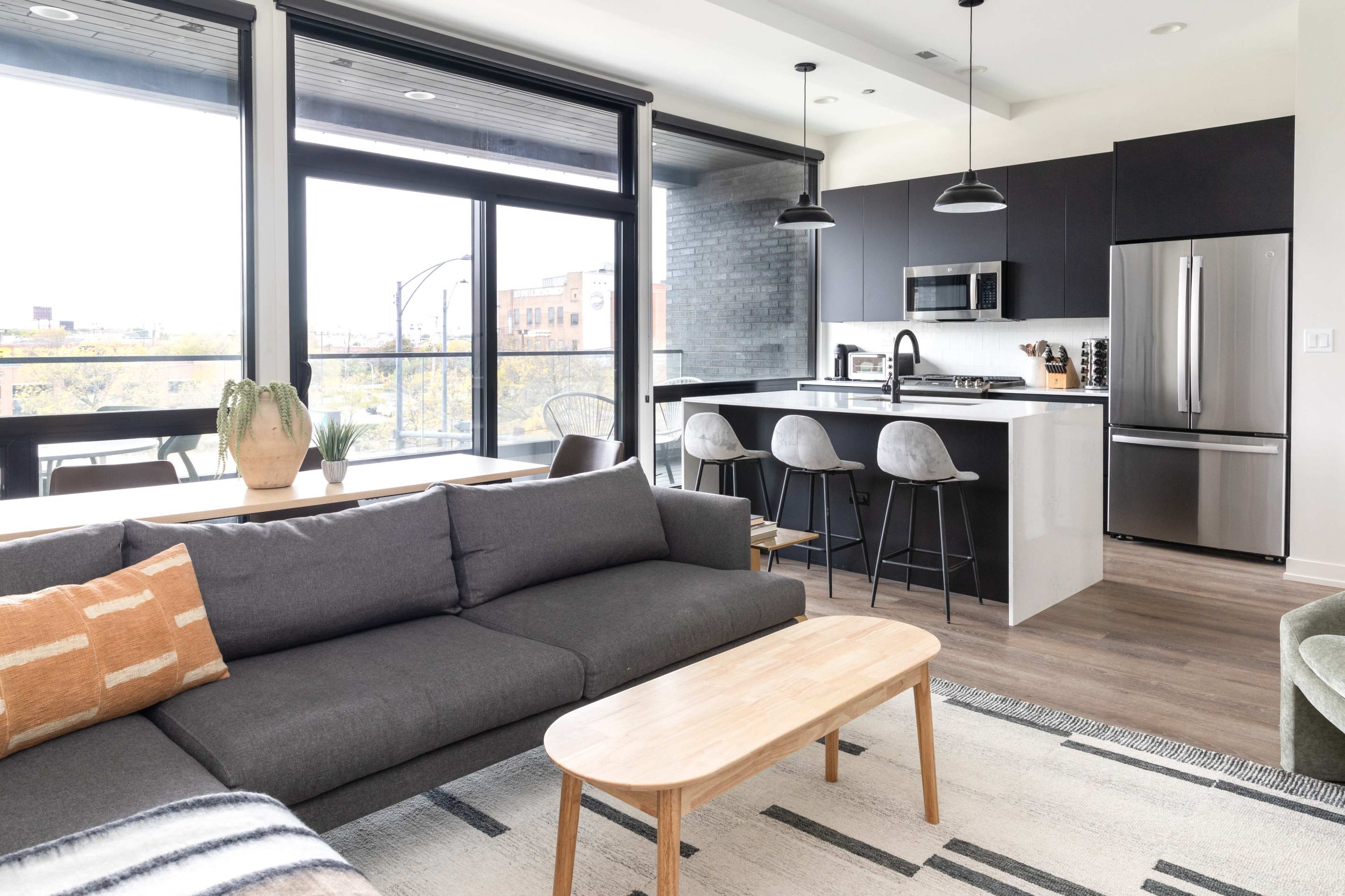 A modern living space featuring a gray sofa, a light wood coffee table, and a kitchen area with dark cabinetry and bar stools.
