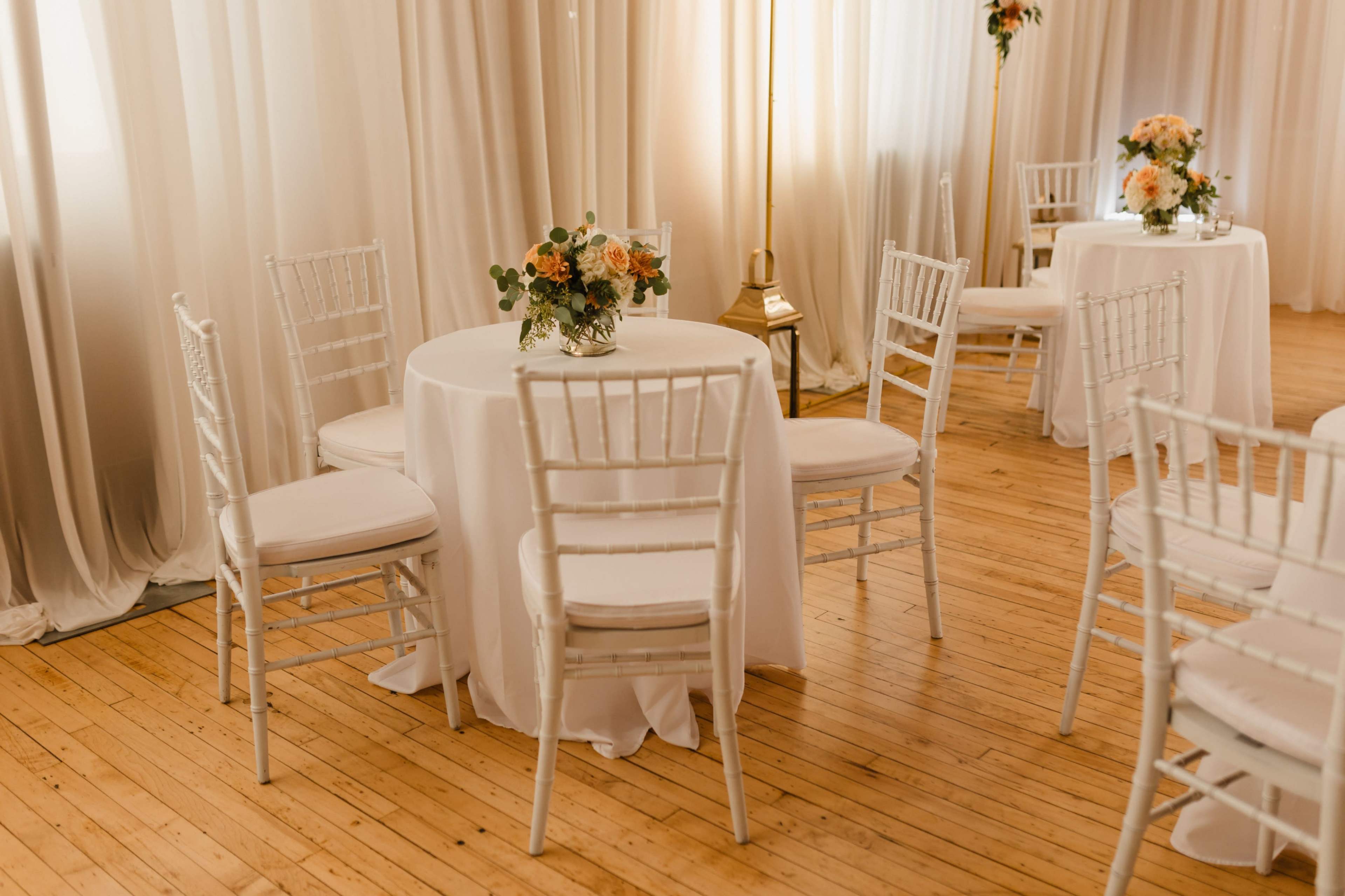 The image shows elegantly set round tables with white tablecloths and floral centerpieces in a softly lit room.