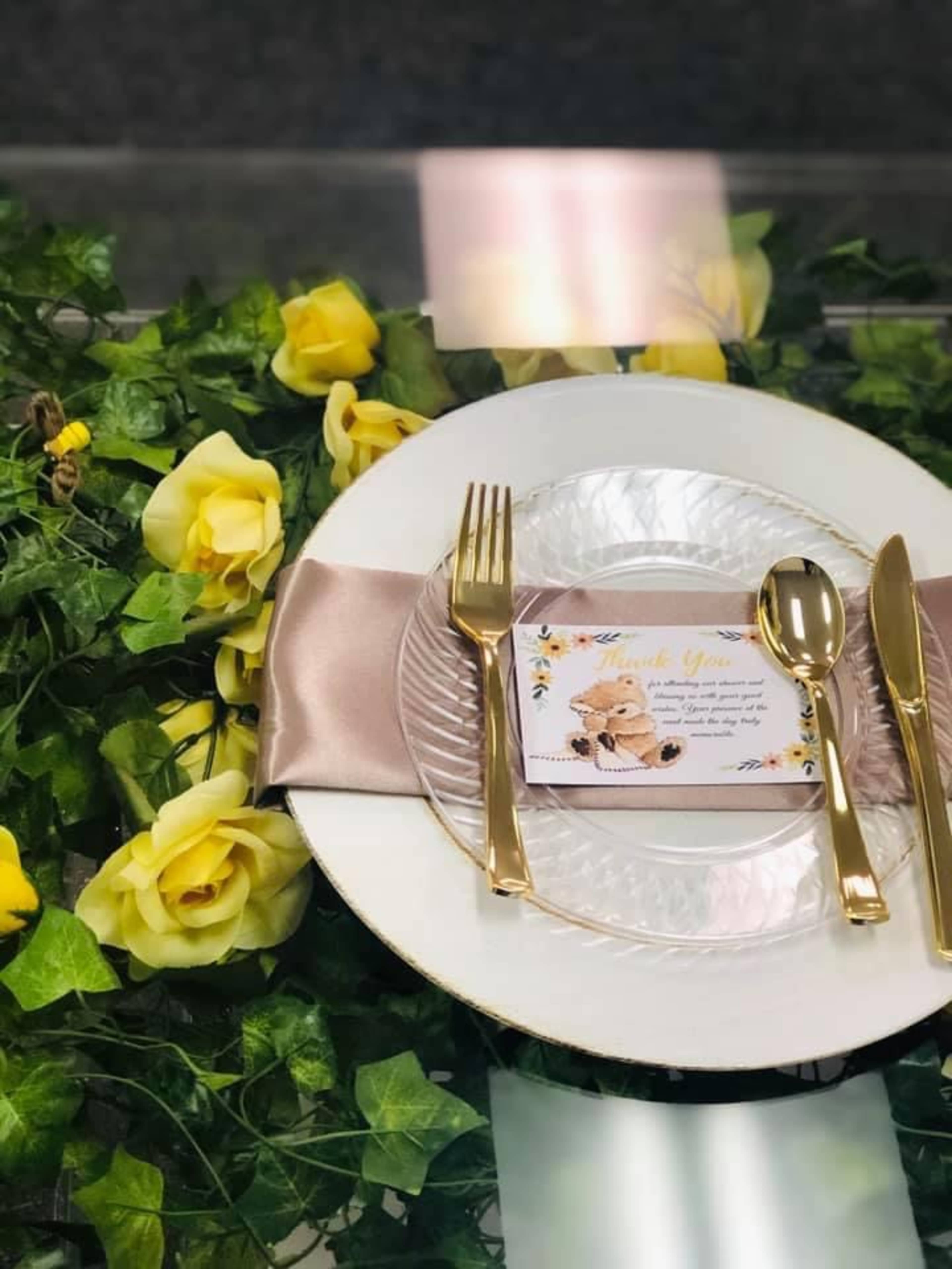 A dining setup features a white plate with golden cutlery, a decorative napkin, and a thank-you card, all arranged on a bed of yellow roses and greenery.