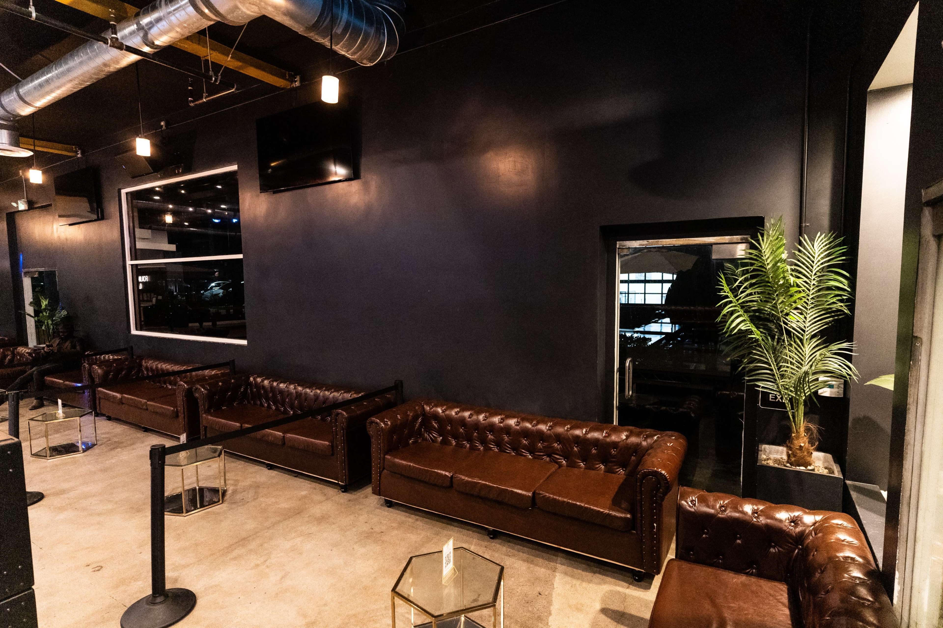 A modern lounge area with brown leather sofas arranged around small glass tables against a dark wall, featuring a potted plant in one corner.