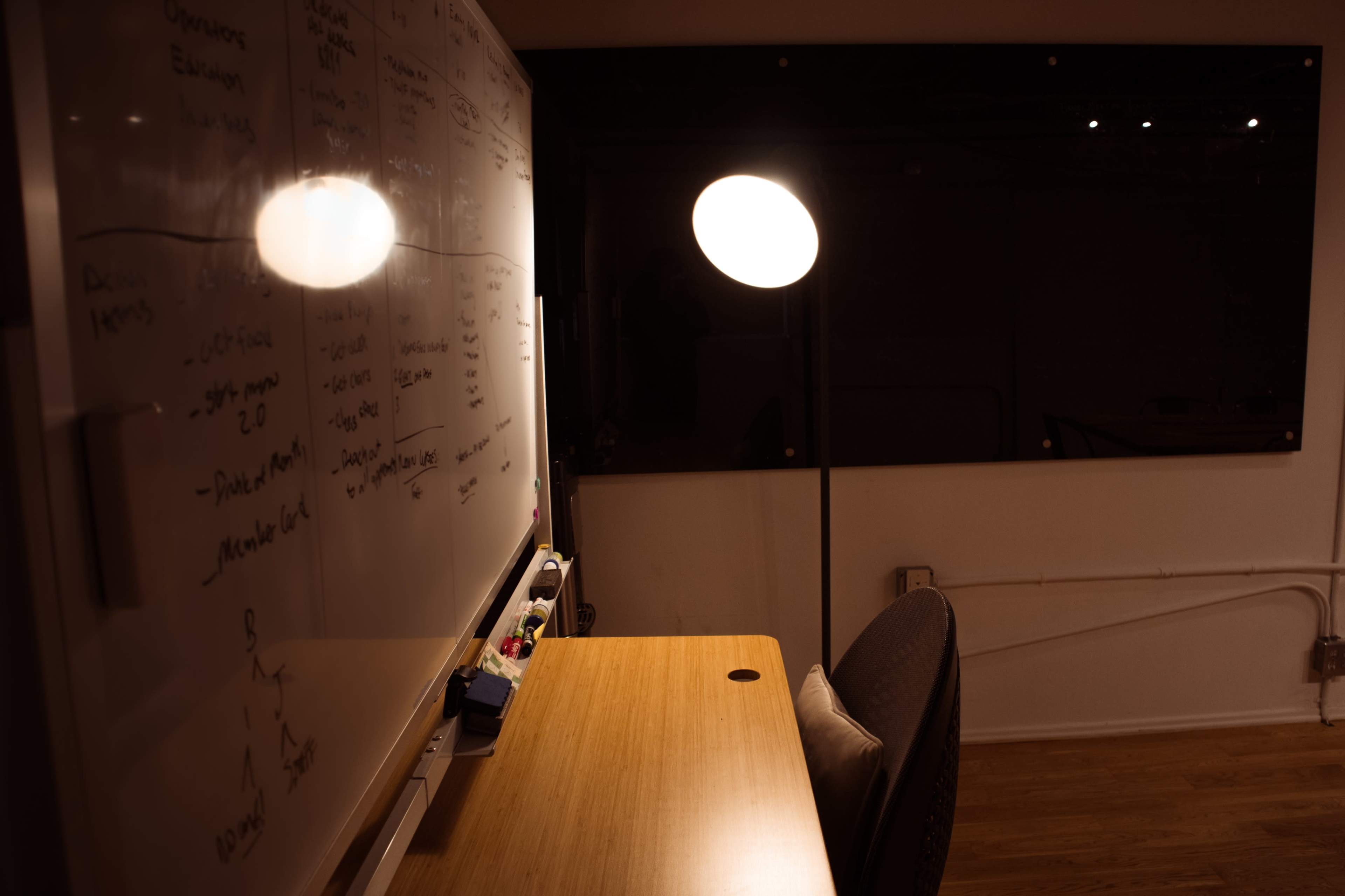 A desk with a chair and a lamp is positioned beside a whiteboard in a dimly lit room.