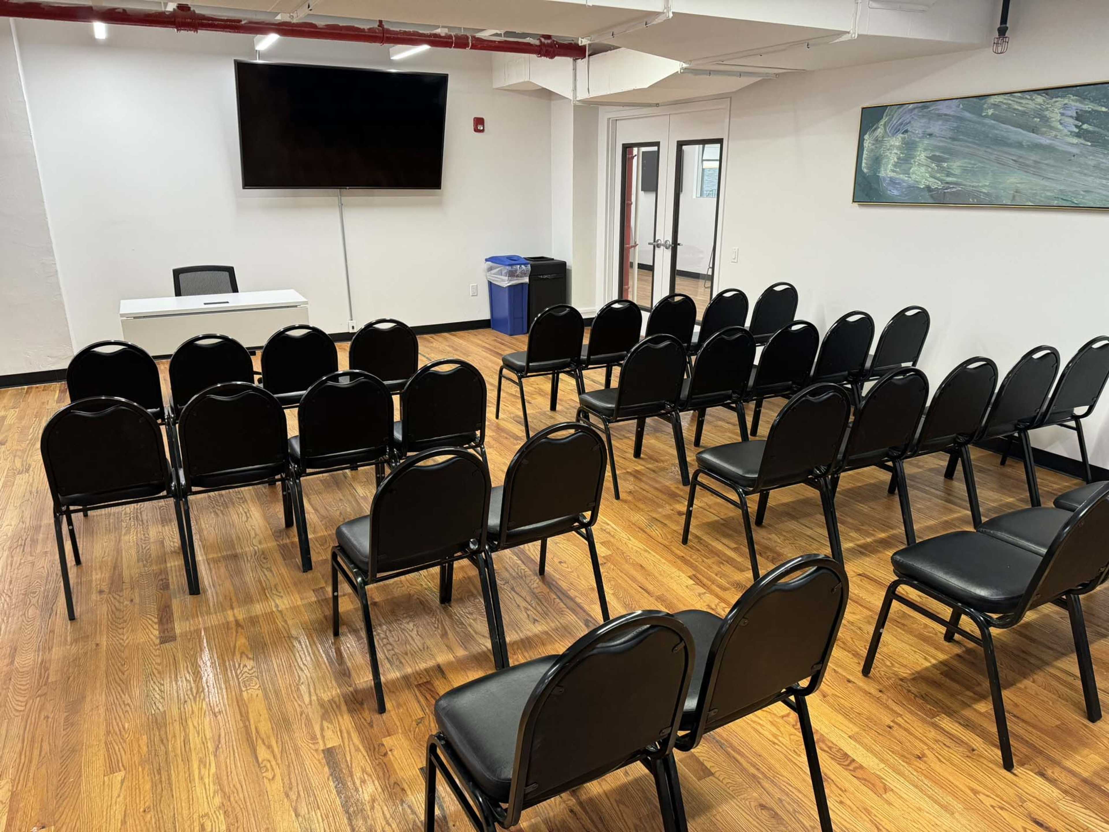 A room is set up with rows of black chairs facing a white table and a large television screen on the wall.