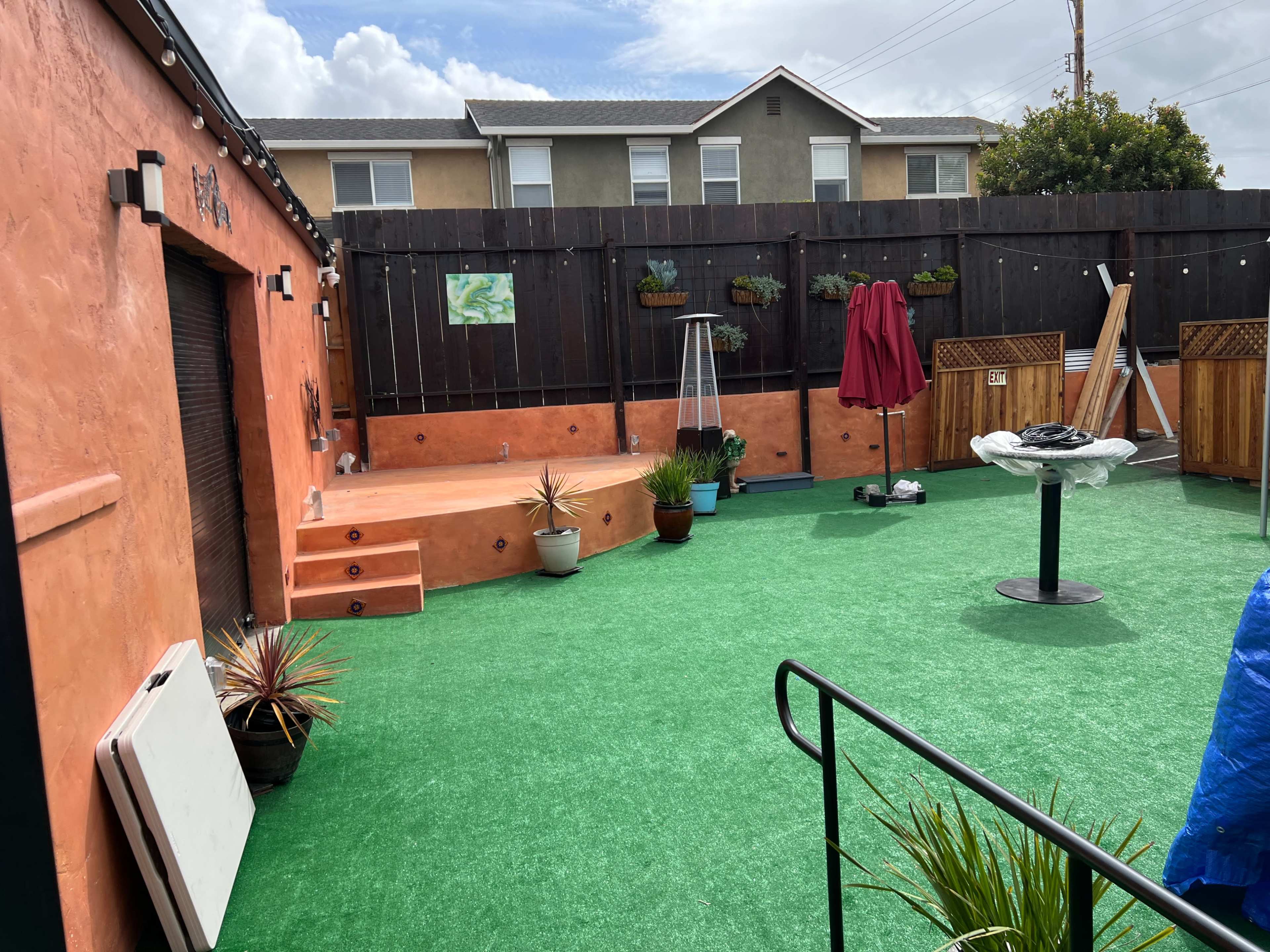 The image shows a fenced outdoor space with artificial grass, a small raised platform, potted plants, and a table under a red umbrella.