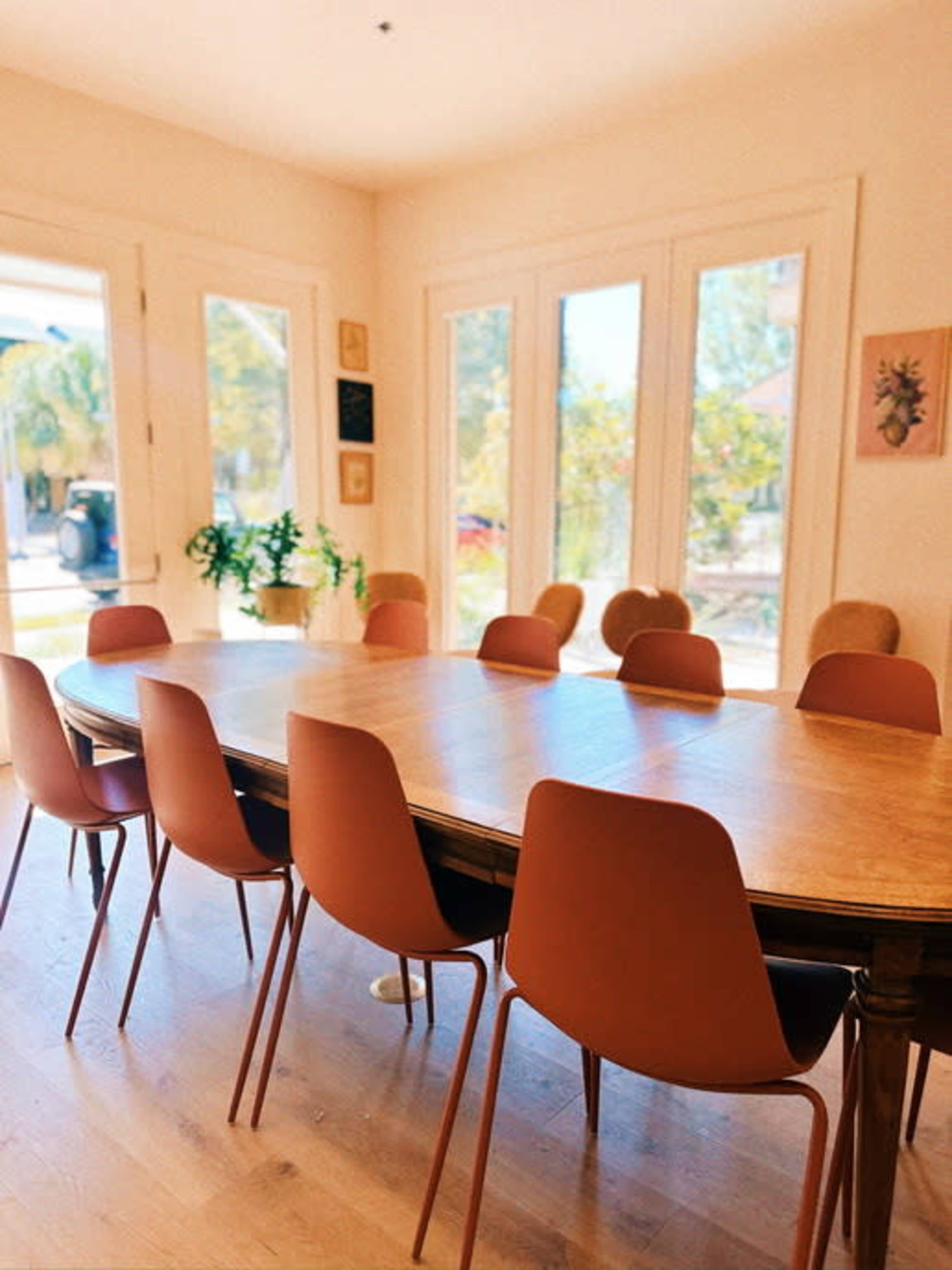 A long wooden dining table is surrounded by several brown chairs in a well-lit room with large windows.