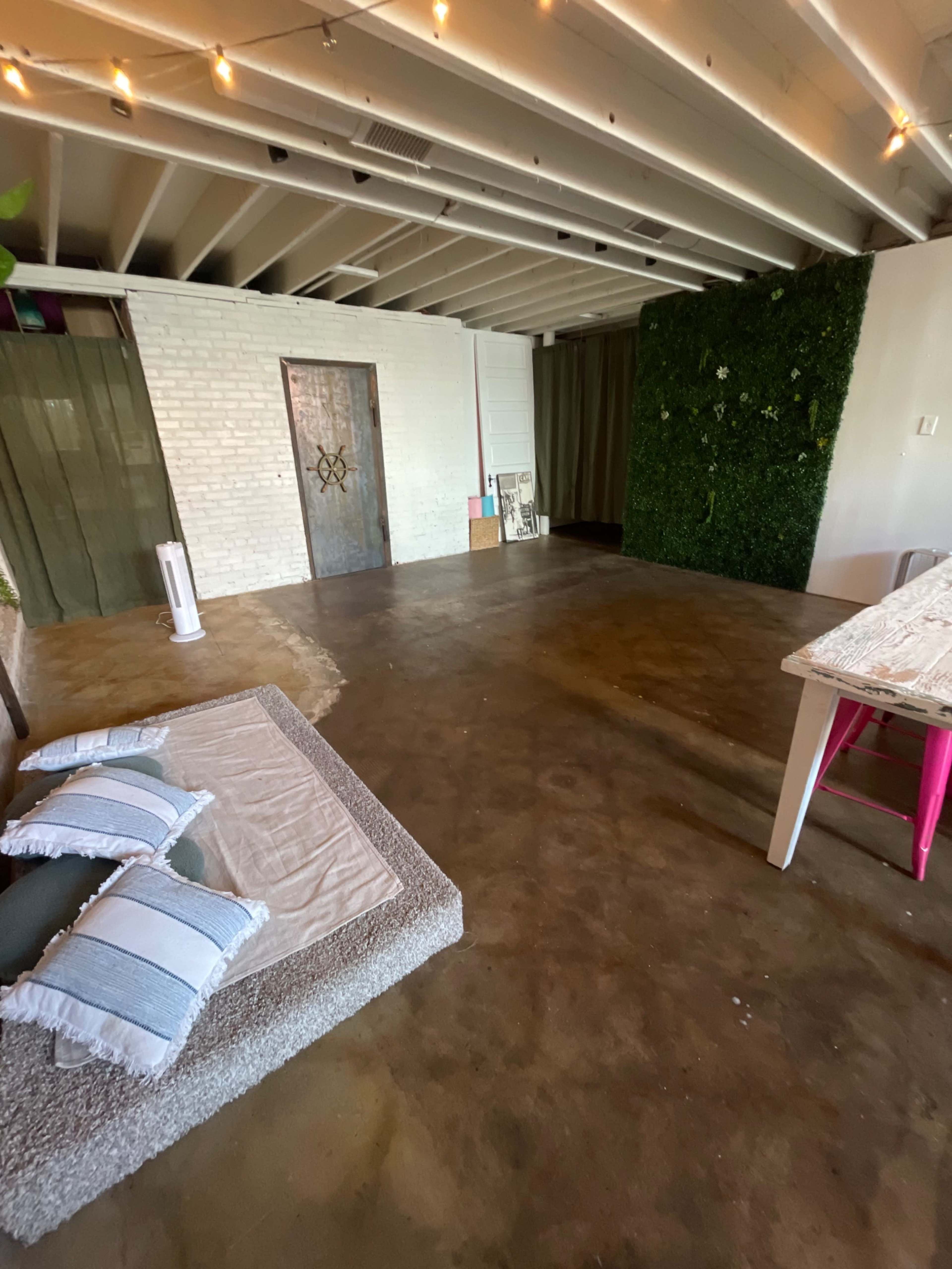 The image shows a spacious room with a concrete floor, a white brick wall, and green foliage on one wall, featuring a sofa with decorative pillows and a small table.