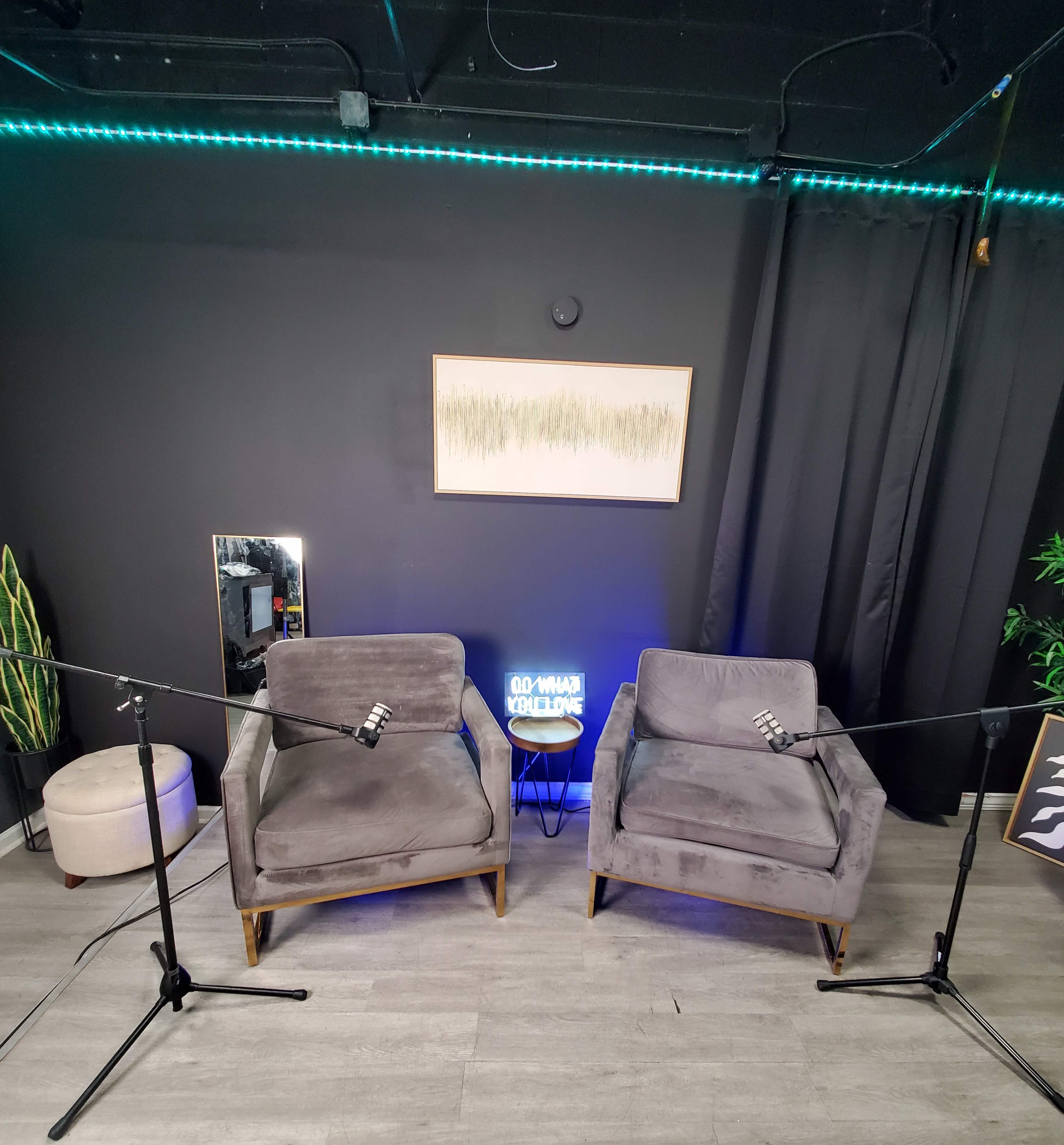 The image shows a modern interview setup with two gray chairs facing each other, flanked by microphone stands, a small table, a decorative mirror, and a green accent light along the wall.
