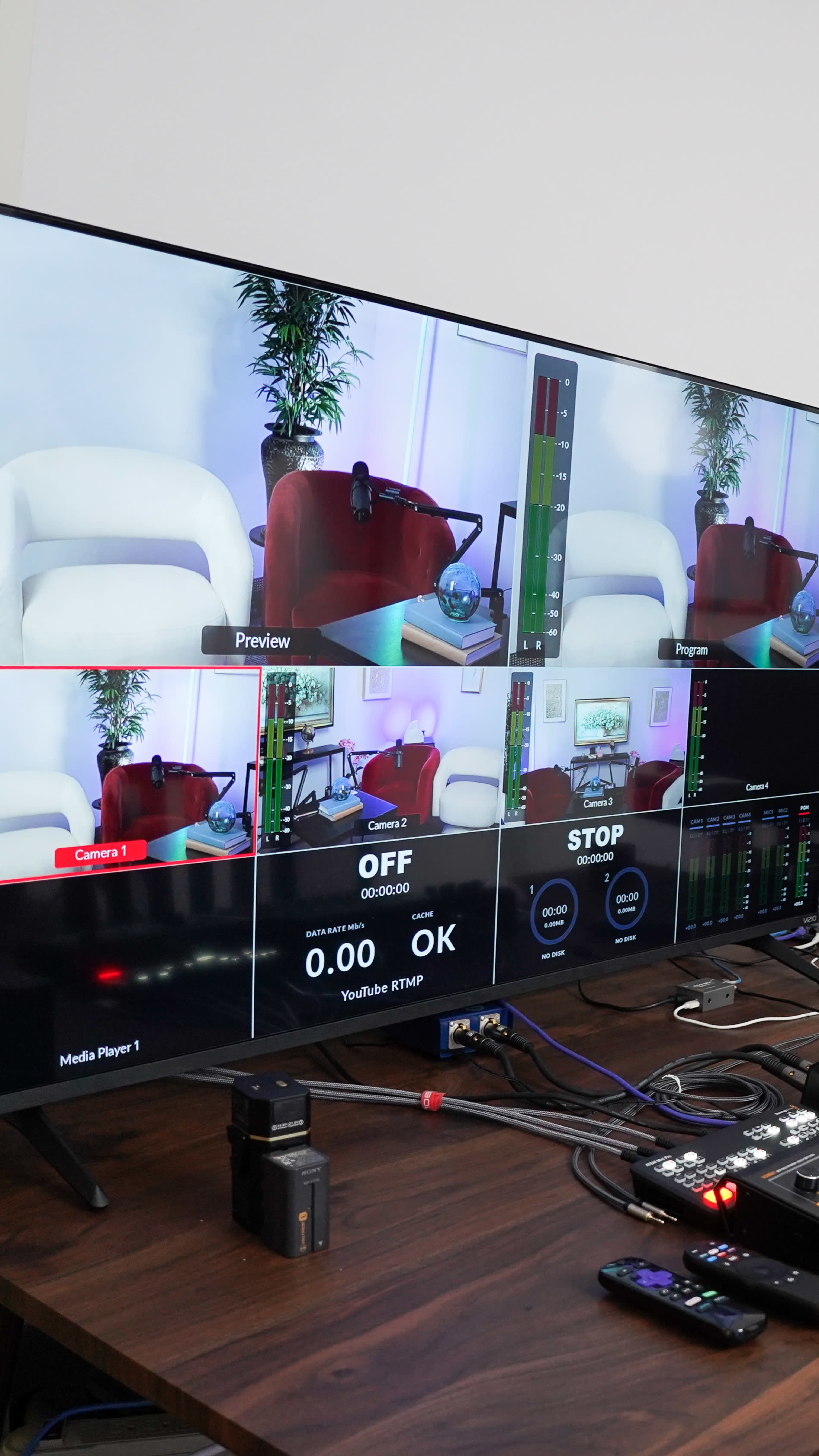 The image shows a large monitor displaying a video production interface with multiple camera feeds, audio levels, and control buttons set on a desk.
