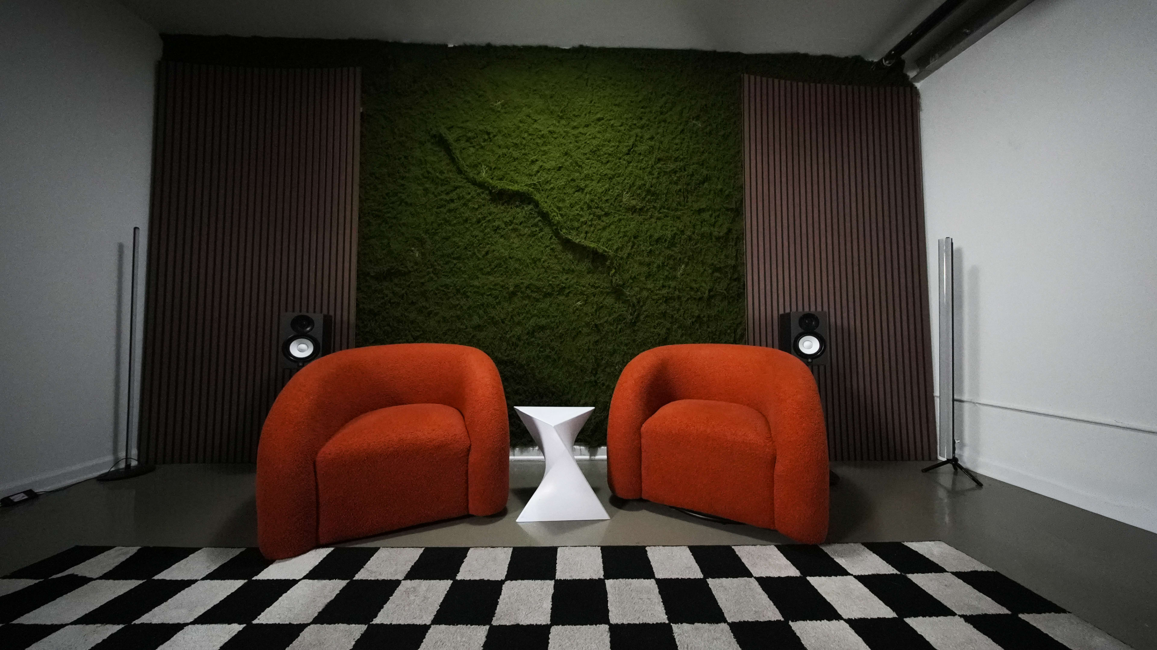 A pair of orange chairs are positioned across from a white geometric table against a backdrop of green moss and wooden panels in a modern interior setting.