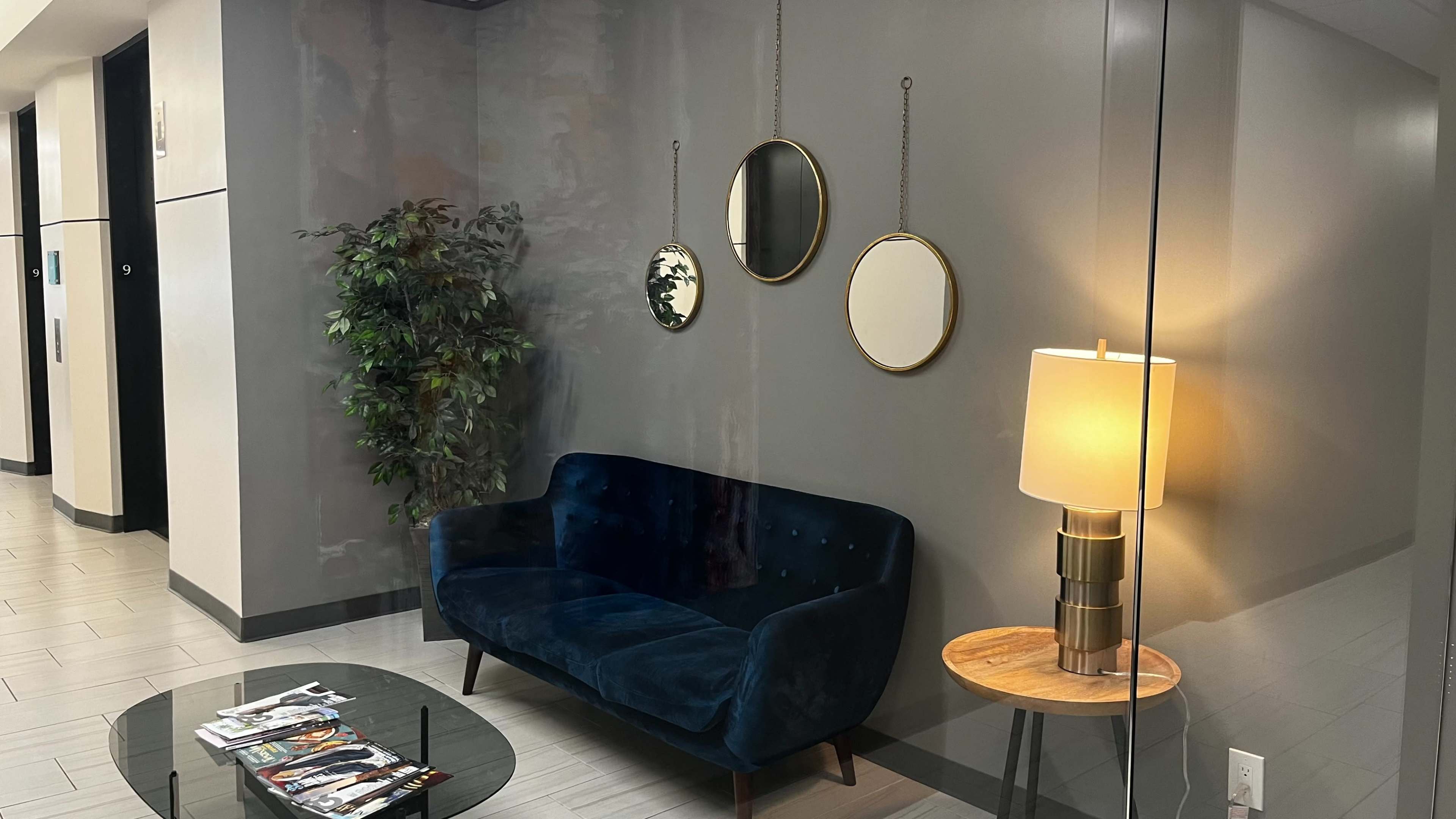 The image shows a modern lobby area with a blue upholstered couch, two round mirrors on the wall, a potted plant, and a lamp on a side table.