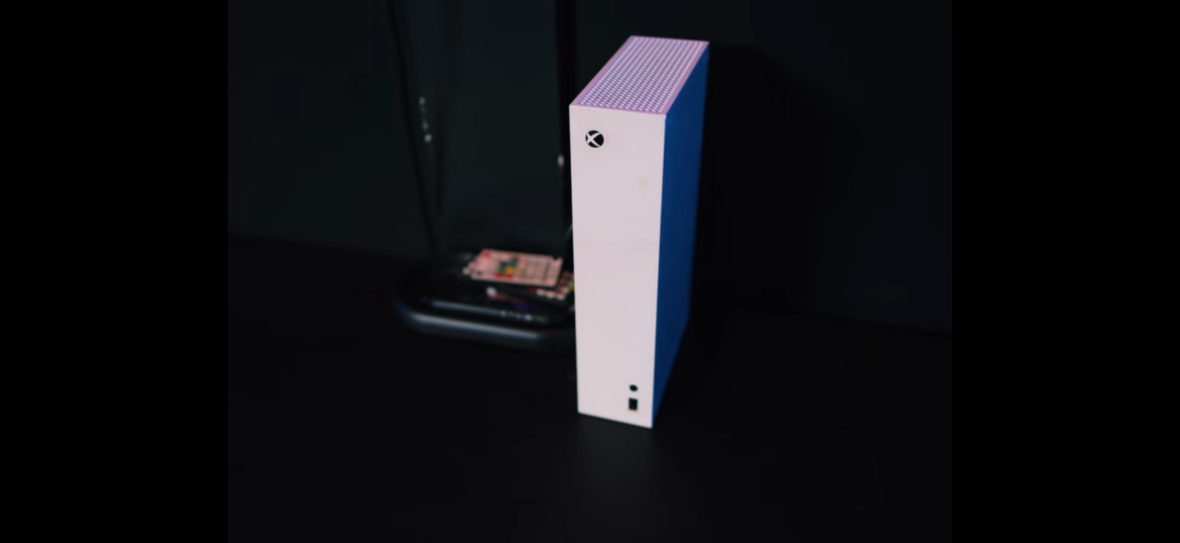 The image shows a white Xbox console positioned vertically on a dark surface, with a transparent case and some electronic components visible in the background.