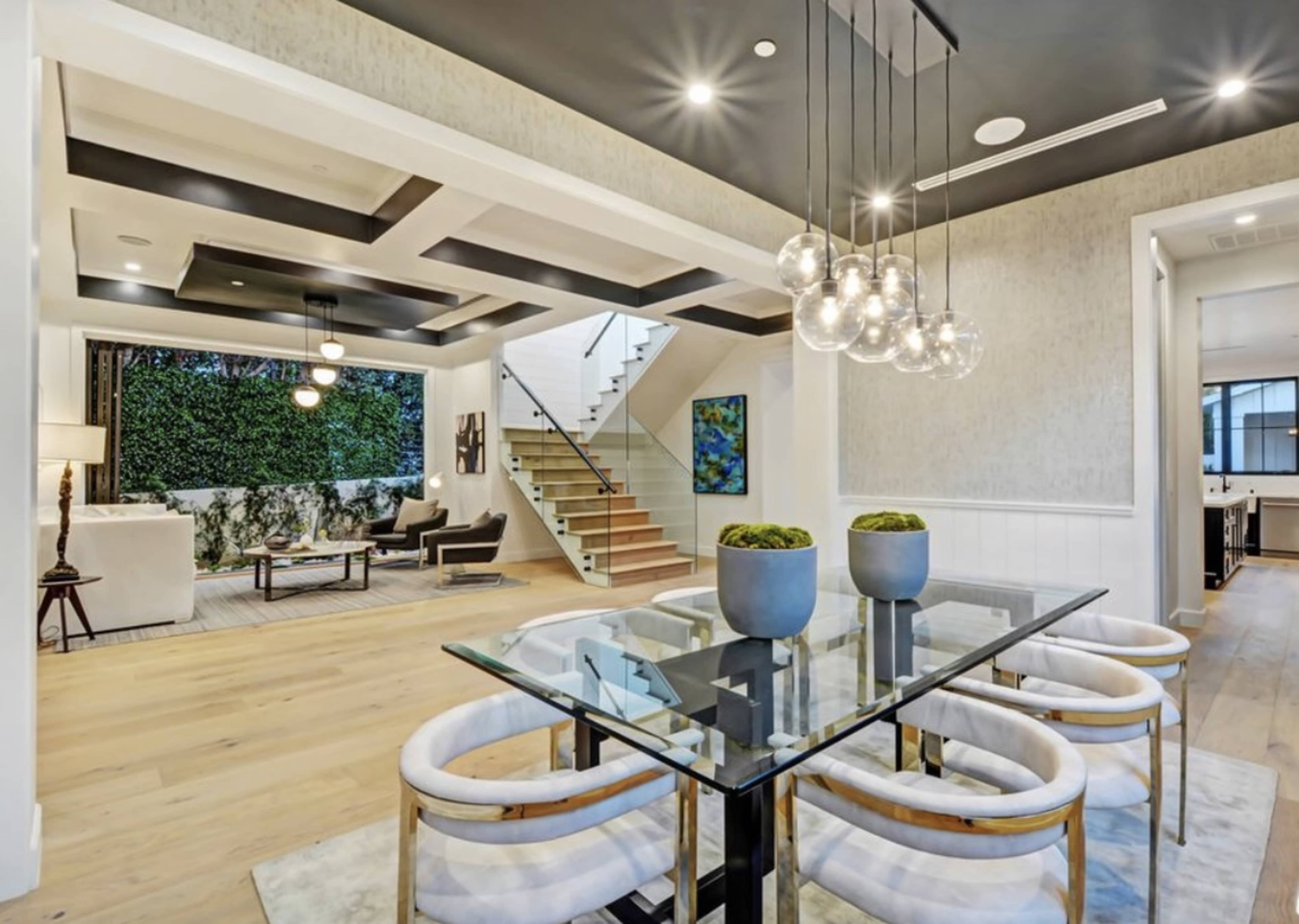 The image shows a modern open-concept living and dining area with a sleek glass dining table, stylish chairs, and a staircase leading to an upper level.