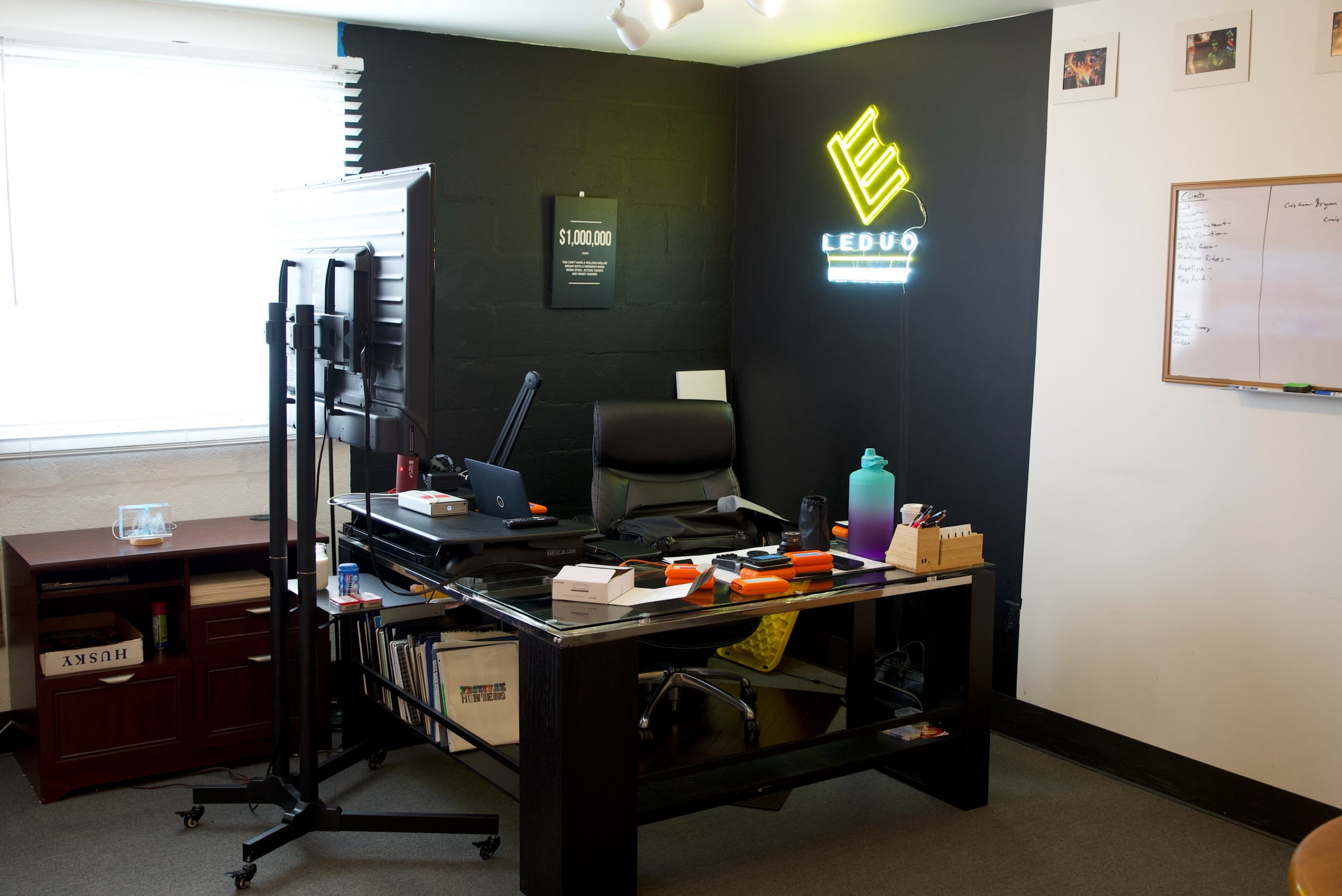 A modern office space features a desk with a computer monitor, chair, various office supplies, and a neon sign on the wall.