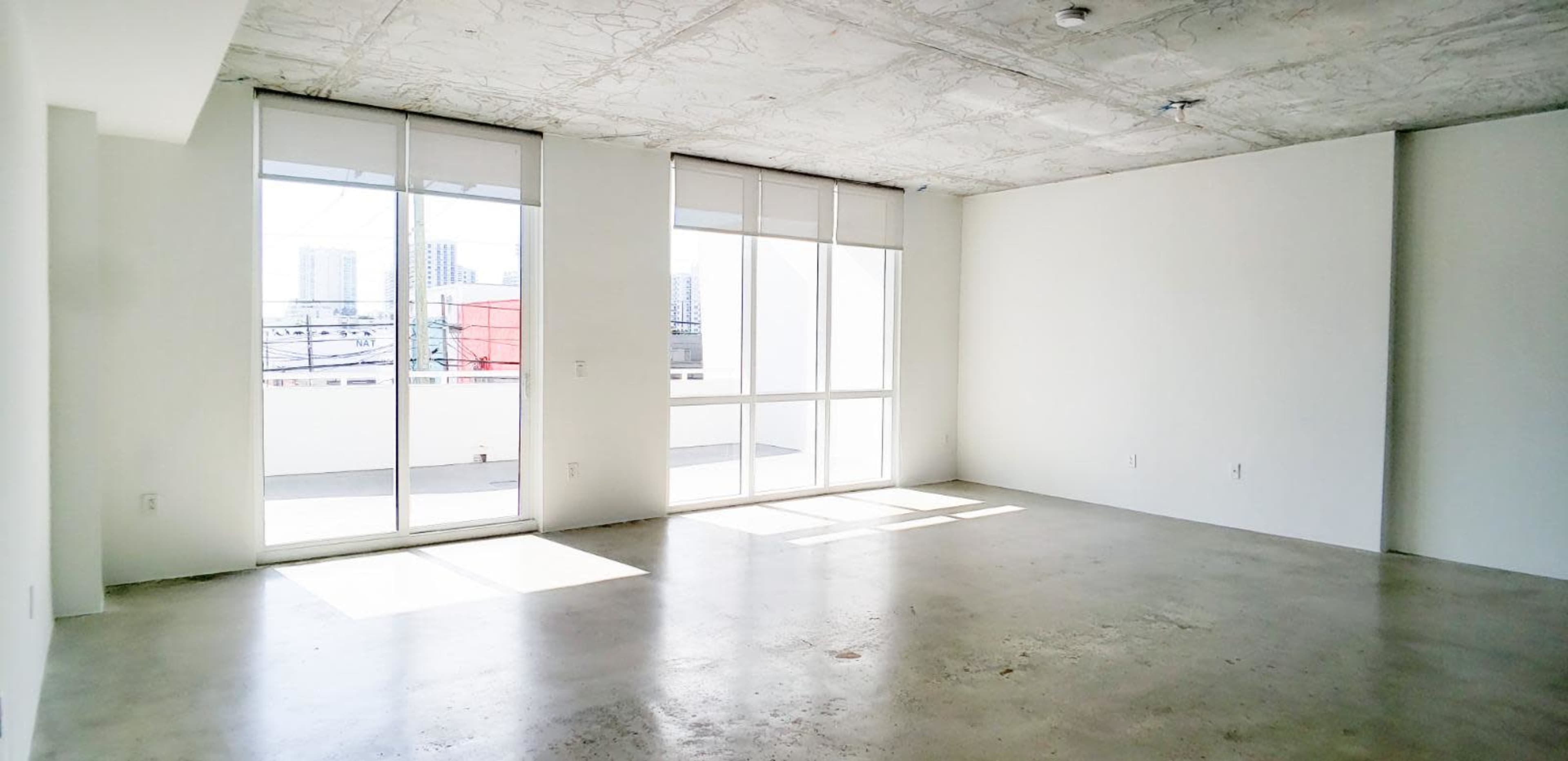 The image shows a spacious, empty room with large windows allowing natural light to enter and a concrete floor.
