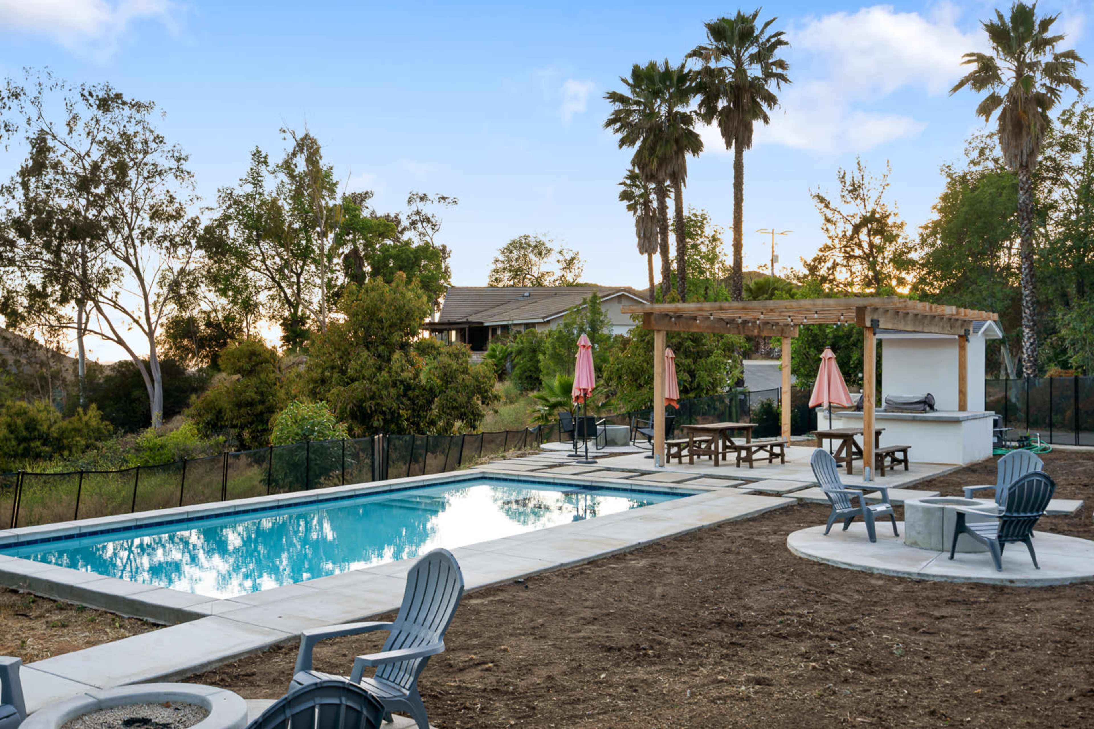 The image shows a swimming pool surrounded by landscaped trees and chairs, with a pergola and barbecue area nearby.