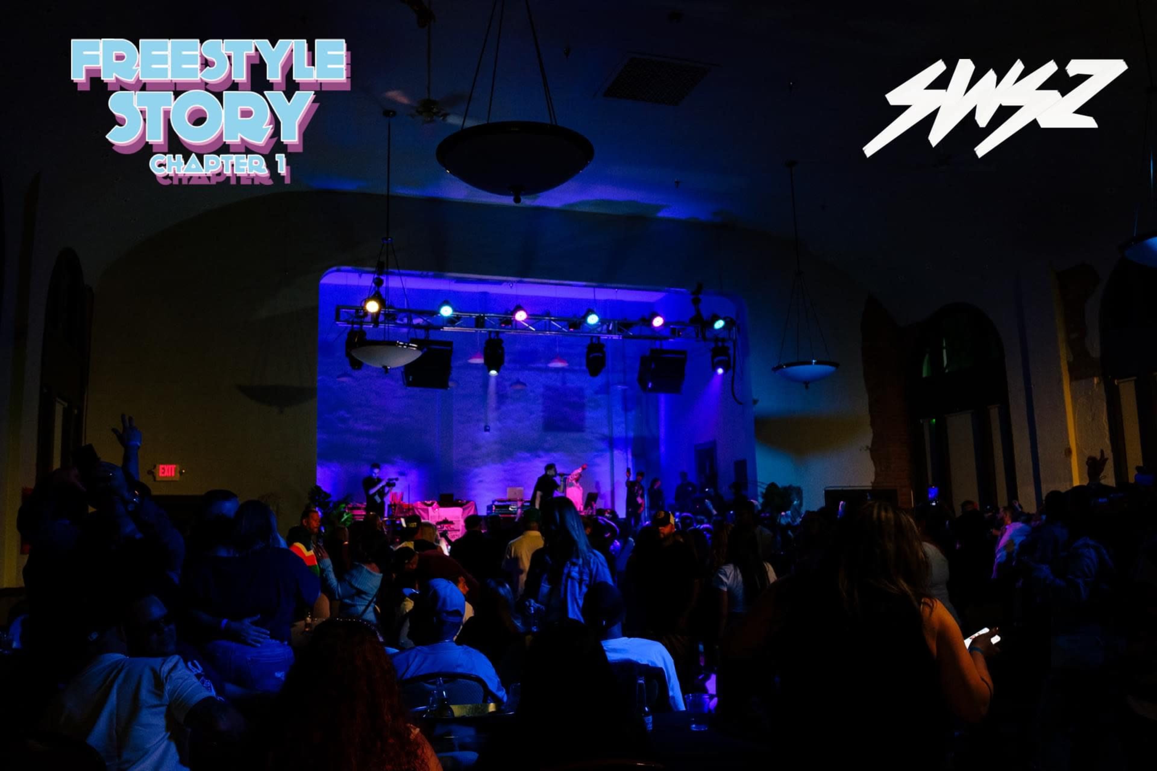 A crowd gathers in a dimly lit room with purple and blue lighting, facing a stage with performers and musical equipment.