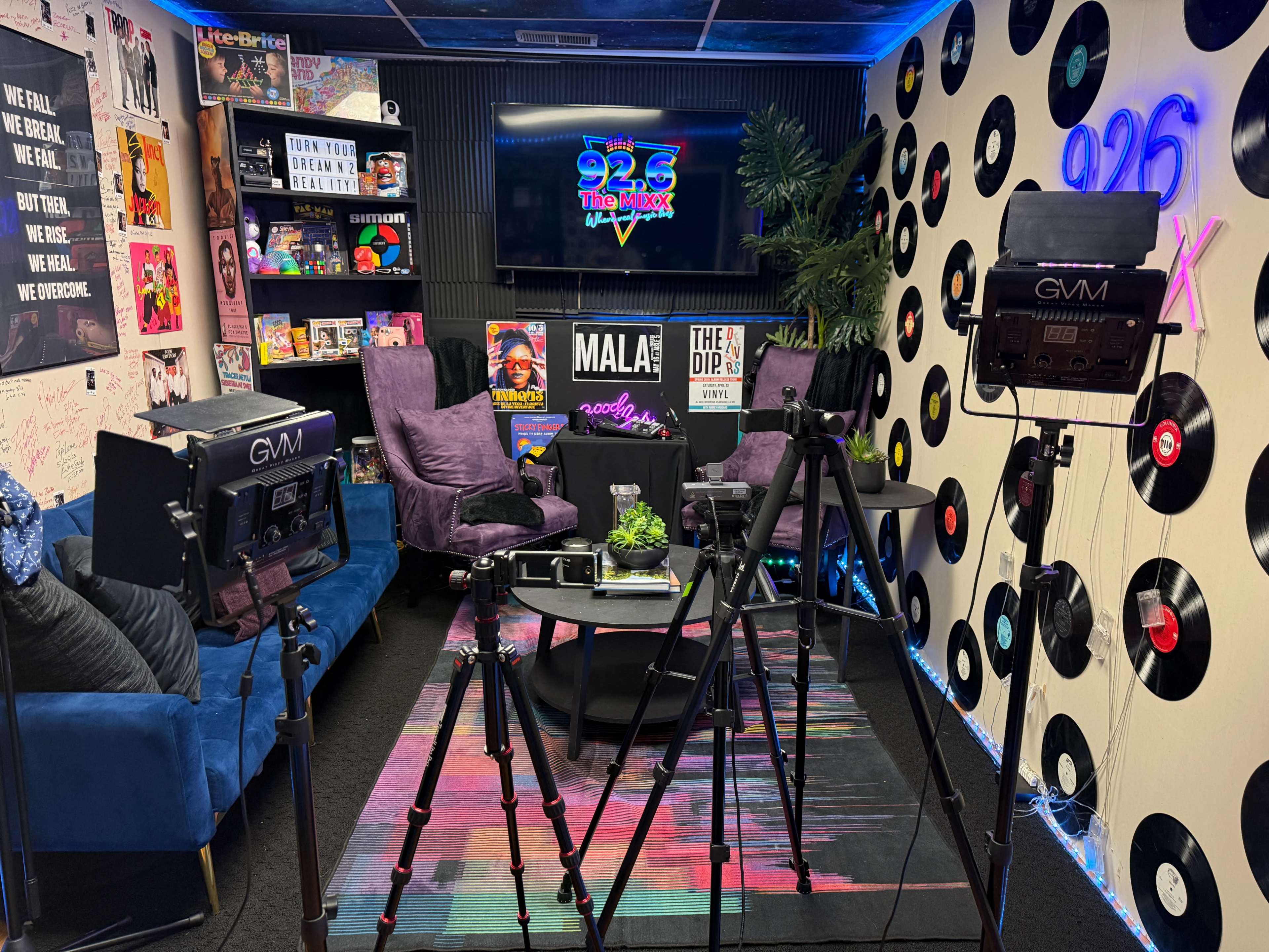 The image shows a brightly decorated studio set with colorful wall decor, featuring a large television screen displaying "92.6 The Mix," surrounded by cameras, lighting equipment, and seating areas with purple chairs.
