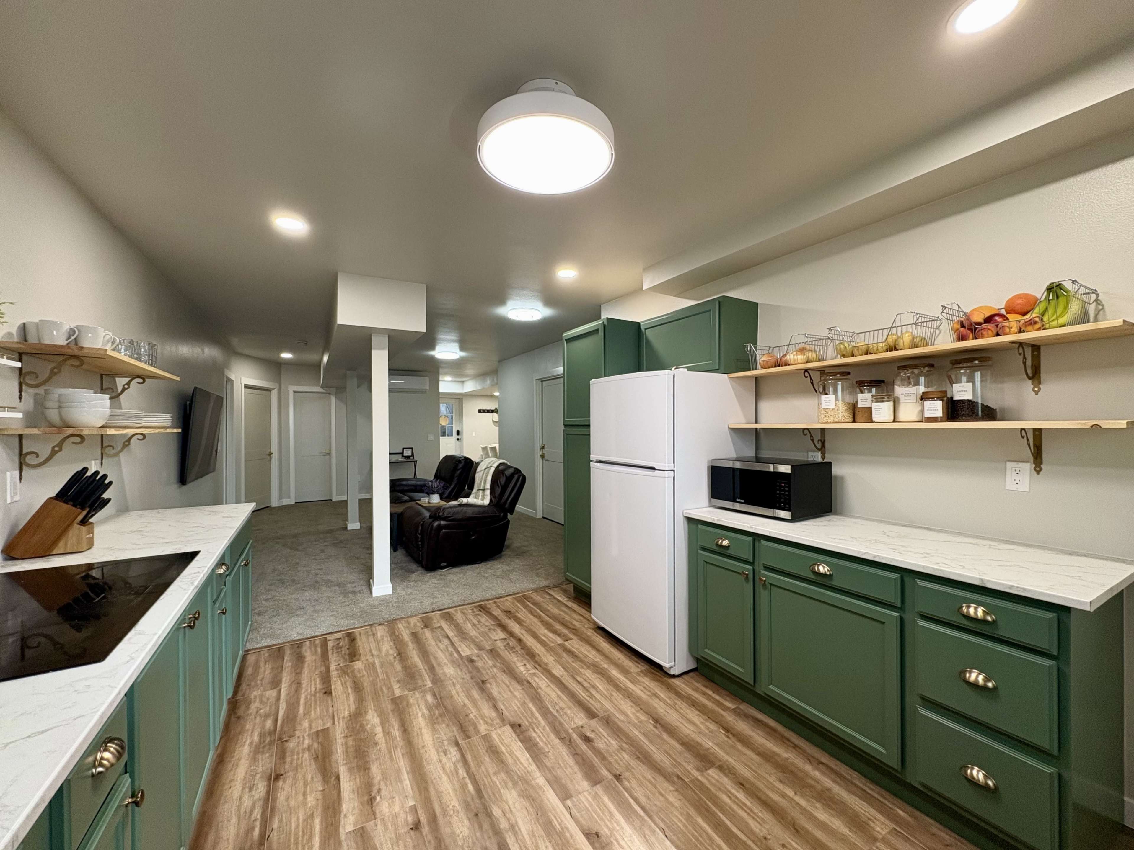 The image shows a modern kitchen with green cabinets, a white refrigerator, and a small dining area beyond, featuring two recliner chairs.