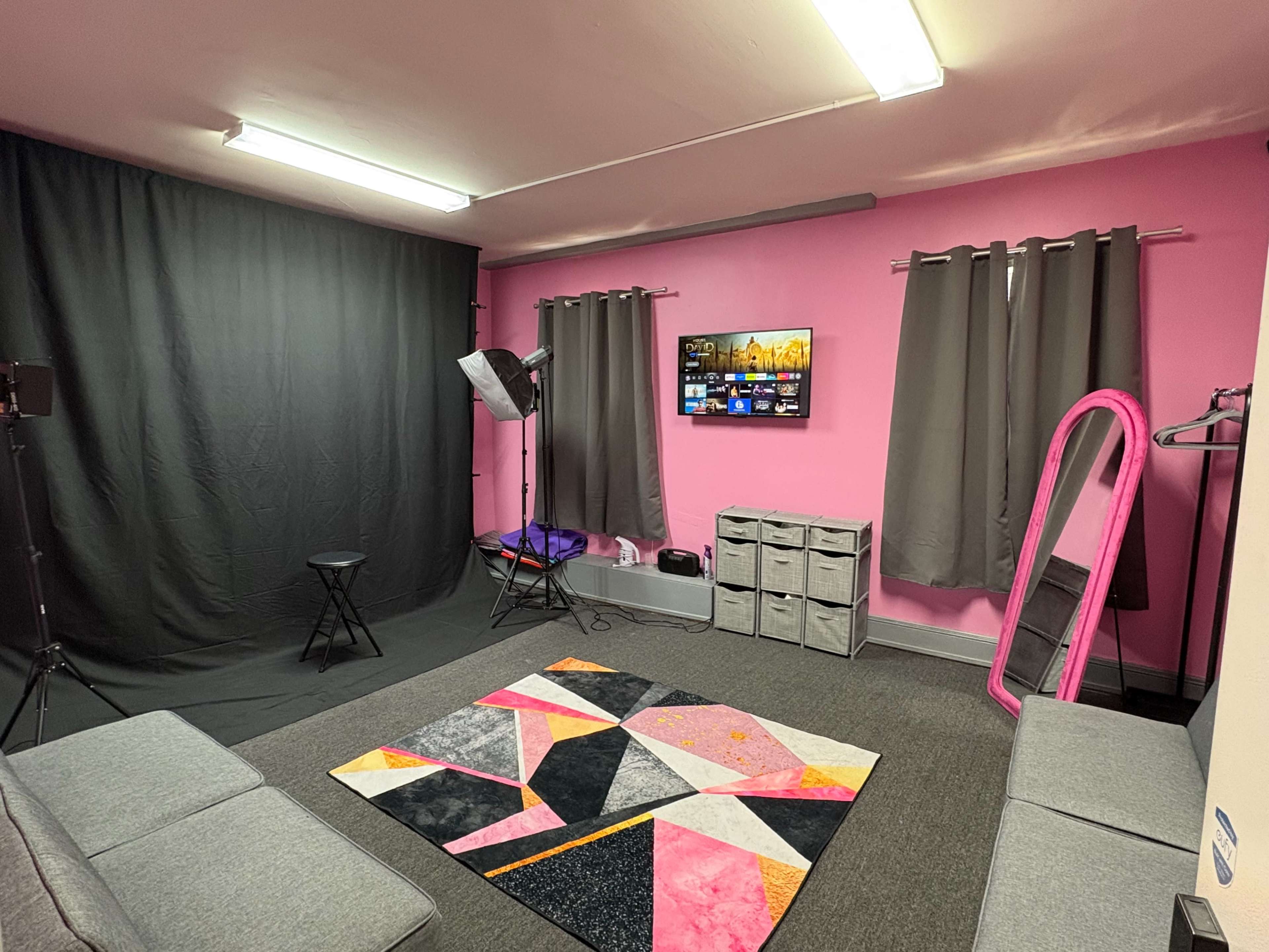 The room features a pink and gray color scheme with a large TV mounted on the wall, black curtains, a mirrored stand, a geometric rug, and seating arrangements along the sides.