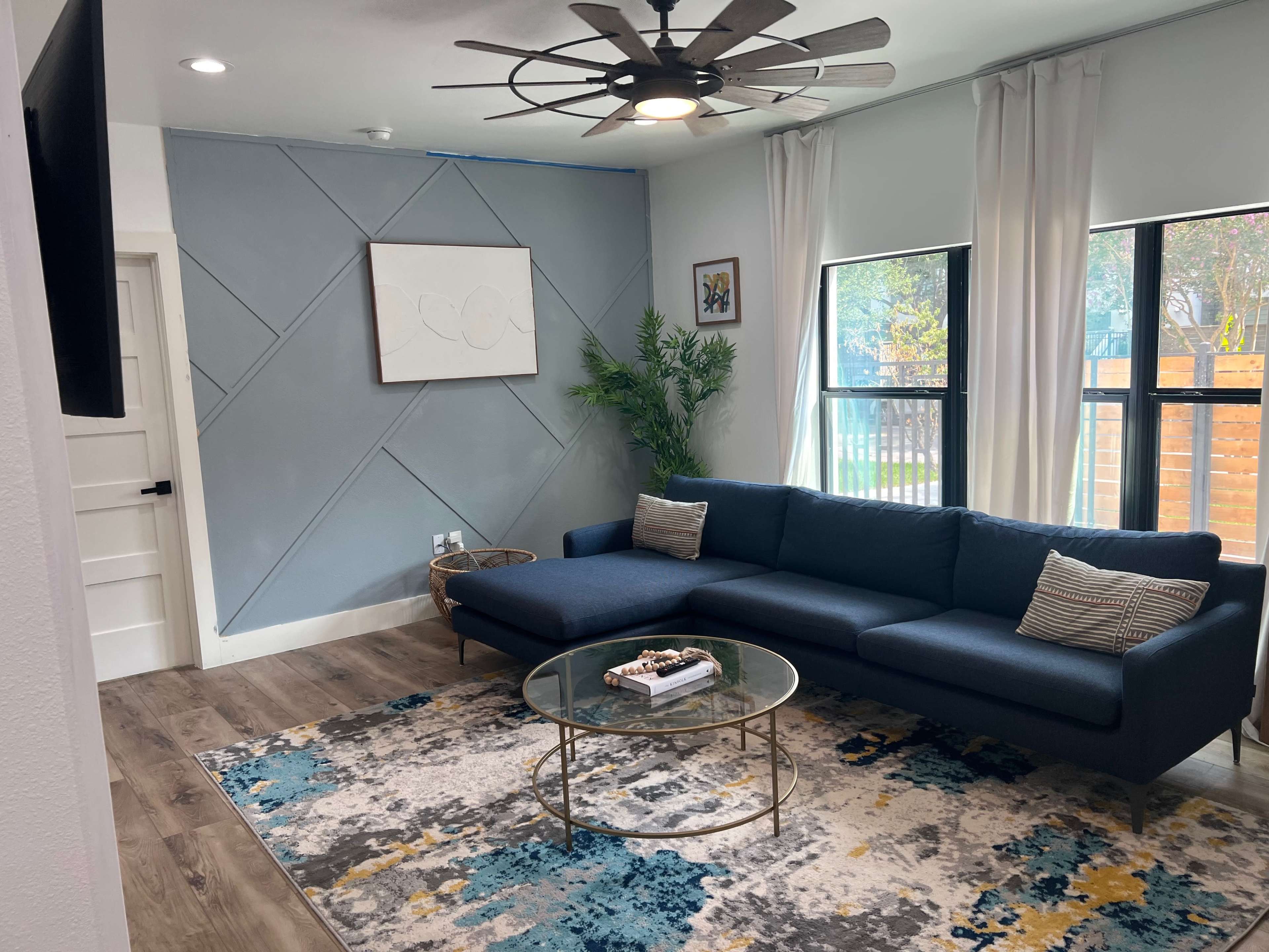 A modern living room featuring a blue sectional sofa, a round glass coffee table, and large windows with white curtains.